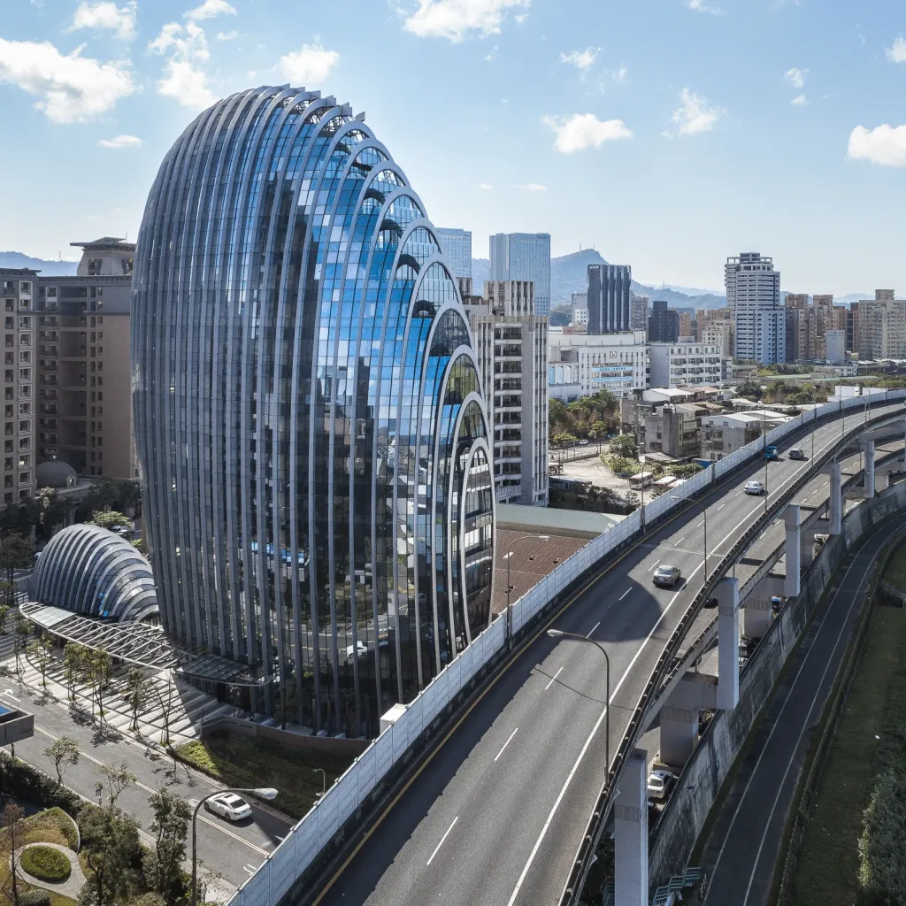 Aedas’ Lè Architecture office tower with rounded form and vertical green façade beside the Jilong River in Taipei. Oblique aerial view of Aedas’s Lè Architecture, an 18‑storey office building with an egg‑shaped massing and curved glass curtain wall. Vertical planted façade bands integrate a green wall system. The tower sits between an elevated highway and river corridor, with a shaded entrance canopy and setback plaza within a subtropical urban context.
