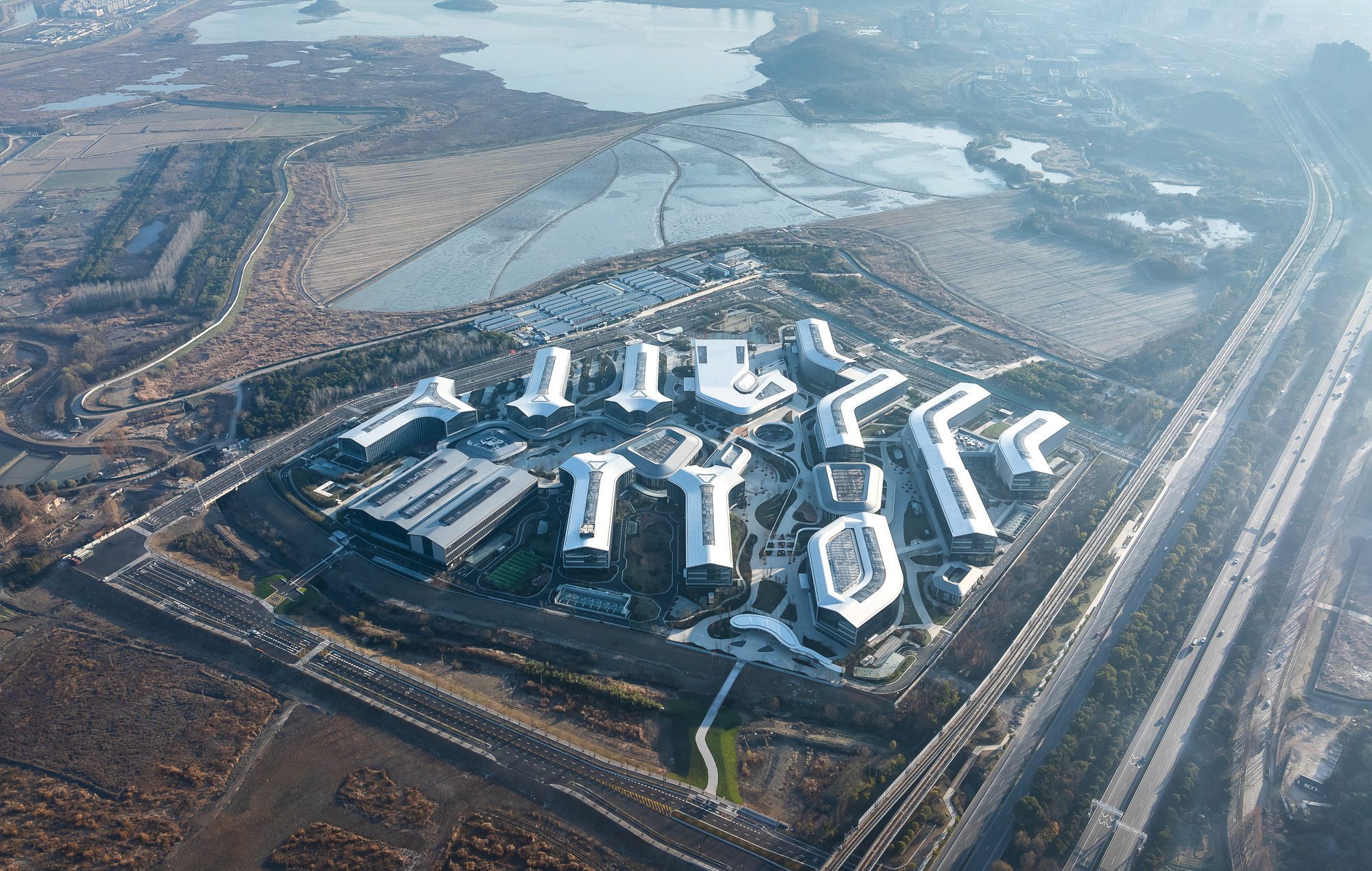 Aedas’ Alibaba Nanhu Future Science Park—research campus with linear laboratory blocks, courtyards and lake‑edge setting in Hangzhou. High‑altitude aerial view of Aedas’ Hangzhou Alibaba Nanhu Future Science Park: a research and innovation campus arranged as parallel, finger‑shaped laboratory and office blocks around landscaped courtyards. Low‑rise volumes with light‑coloured roofs embed within lakeside terrain and infrastructure corridors—supporting campus planning, daylight‑oriented layouts and landscape‑integrated R&D environments.