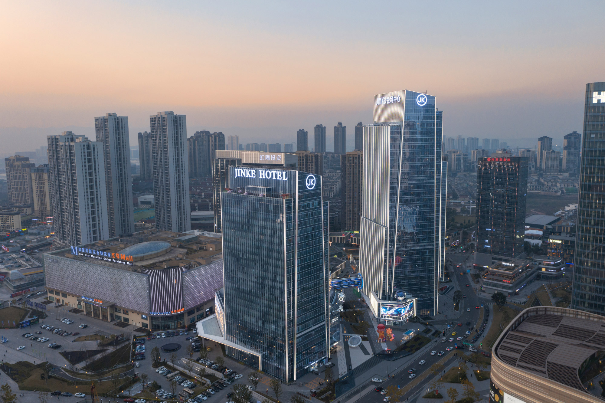 Aedas’ Jinke Group Headquarters in Chongqing with stepped glass towers and gold‑toned curtain wall massing. Aerial dusk view of the Aedas‑designed Jinke Group Headquarters in Chongqing, showing stepped high‑rise office and hotel towers clad in high‑performance glass curtain walls and aluminium panels. The towers anchor a mixed‑use podium with retail frontage and internal circulation corridors, set within a dense urban district shaped by hilly topography and arterial road networks.