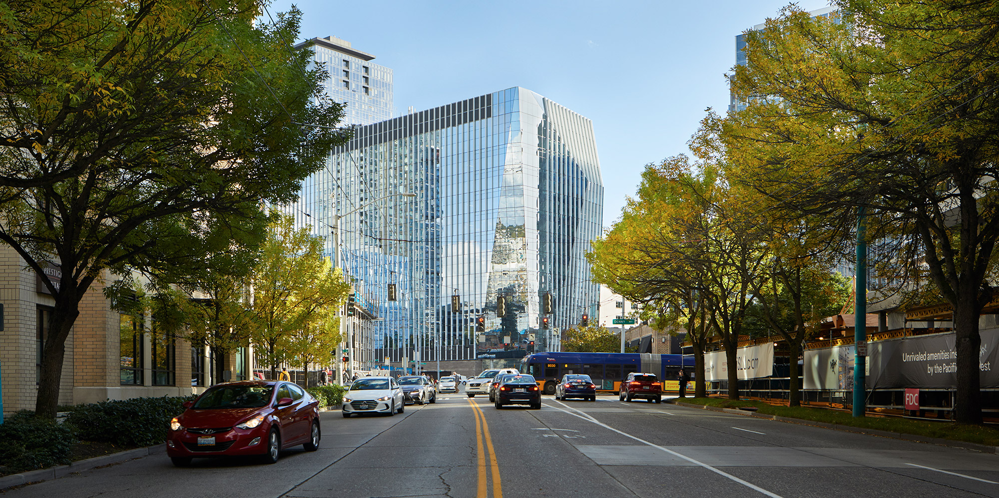 Aedas’ Building Cure immunotherapy research tower in Seattle with high‑performance glazed façade and urban street interface. Street-level view of Aedas-designed Building Cure in Seattle’s Denny Triangle, showing a mid-rise research tower with floor-to-ceiling glazed curtain wall and vertical façade modulation. The building addresses an active urban street with integrated ground-level public space, framed by mature street trees and adjacent mixed-use development within a temperate Pacific Northwest climate.