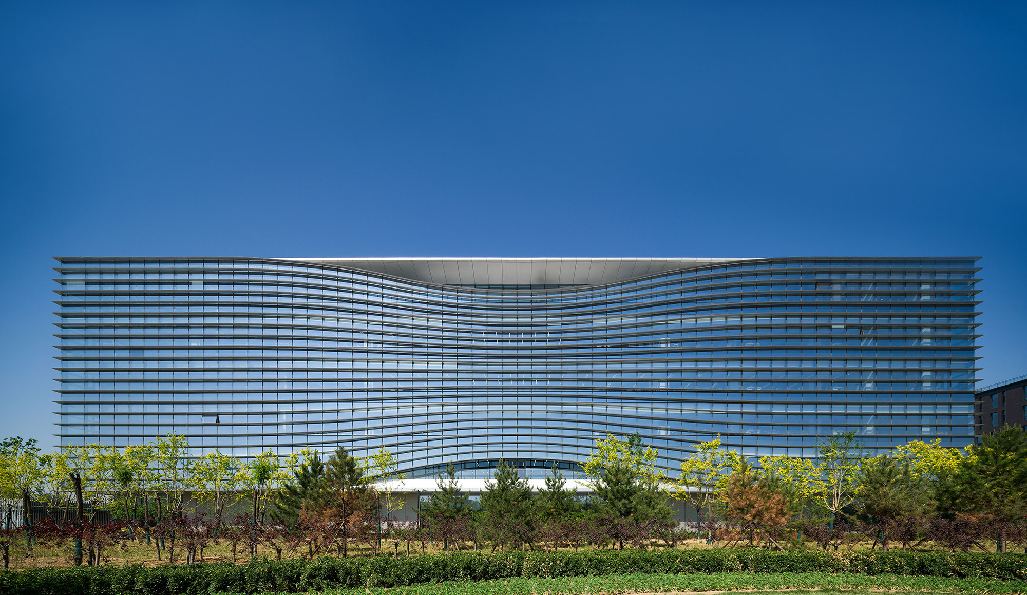 Aedas’ Sina Plaza headquarters with rippling glass façade and horizontal solar fins in Zhongguancun, Beijing. Front elevation of Aedas’s Sina Plaza showing a low‑rise headquarters building with a concave, wave‑like façade. The envelope uses floor‑to‑ceiling glazed curtain wall panels articulated by continuous horizontal shading fins. A recessed central roof opening marks the main atrium, while landscaped foreground planting frames the campus setting in Beijing’s temperate climate.