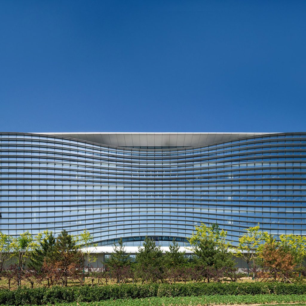 Aedas’ Sina Plaza headquarters with rippling glass façade and horizontal solar fins in Zhongguancun, Beijing. Front elevation of Aedas’s Sina Plaza showing a low‑rise headquarters building with a concave, wave‑like façade. The envelope uses floor‑to‑ceiling glazed curtain wall panels articulated by continuous horizontal shading fins. A recessed central roof opening marks the main atrium, while landscaped foreground planting frames the campus setting in Beijing’s temperate climate.