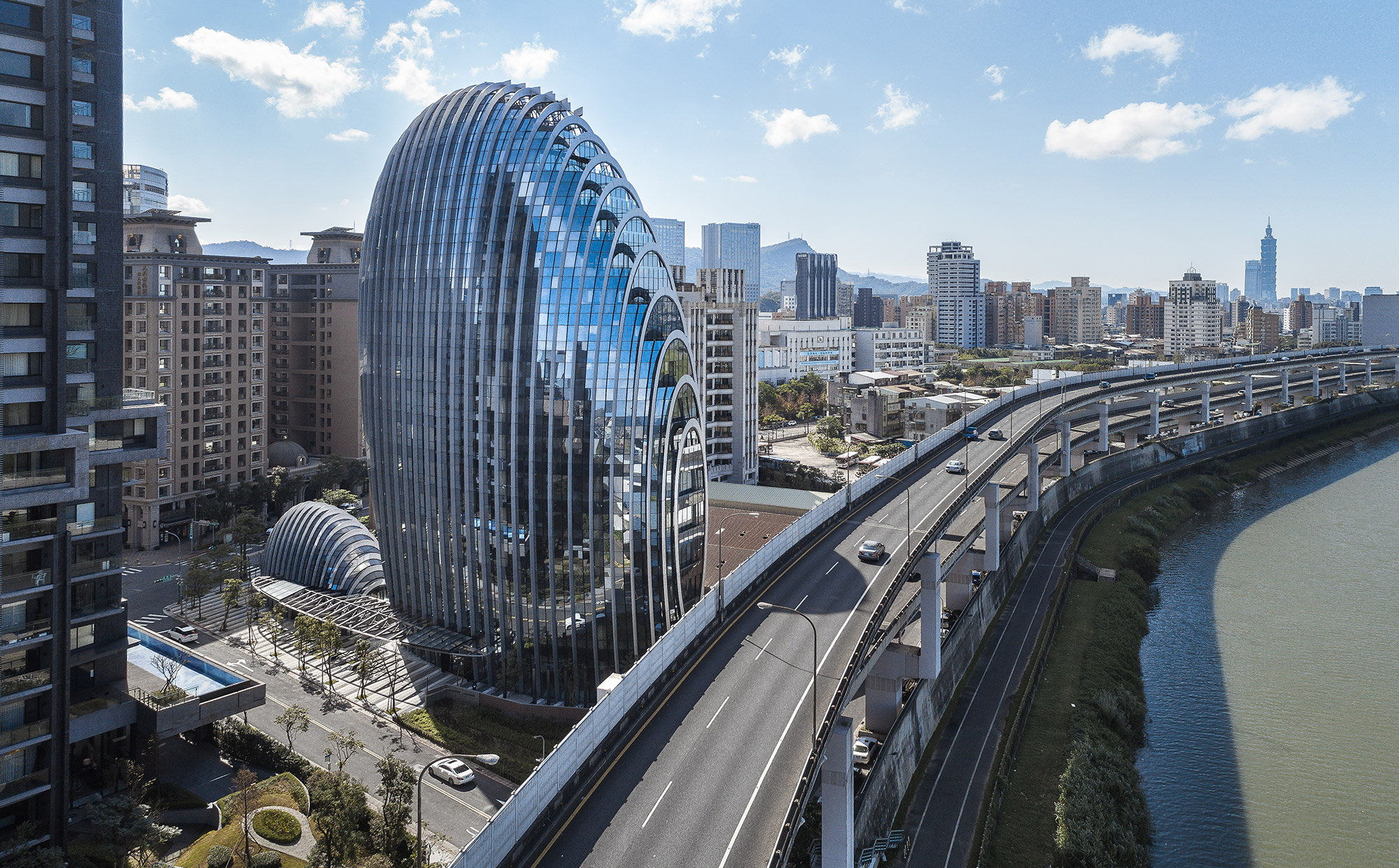 Aedas’ Lè Architecture office tower with rounded form and vertical green façade beside the Jilong River in Taipei. Oblique aerial view of Aedas’s Lè Architecture, an 18‑storey office building with an egg‑shaped massing and curved glass curtain wall. Vertical planted façade bands integrate a green wall system. The tower sits between an elevated highway and river corridor, with a shaded entrance canopy and setback plaza within a subtropical urban context.