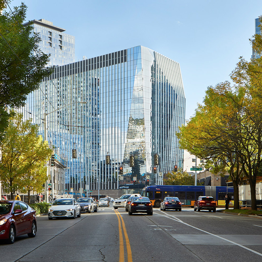 Aedas’ Building Cure immunotherapy research tower in Seattle with high‑performance glazed façade and urban street interface. Street-level view of Aedas-designed Building Cure in Seattle’s Denny Triangle, showing a mid-rise research tower with floor-to-ceiling glazed curtain wall and vertical façade modulation. The building addresses an active urban street with integrated ground-level public space, framed by mature street trees and adjacent mixed-use development within a temperate Pacific Northwest climate.