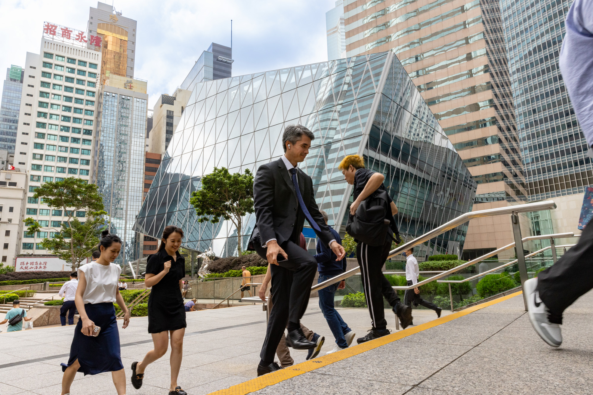 Aedas’ The Forum, Central Hong Kong—five-storey office with faceted glass curtain wall and urban public forecourt. Street-level view of Aedas’ The Forum in Central, Hong Kong: a five-storey office building with faceted, diamond-pattern glass curtain wall reflecting the dense CBD skyline. A stepped public forecourt and integrated planting connect pedestrian movement to surrounding streets. The image evidences high-performance glazing, daylight optimisation and urban infill redevelopment.