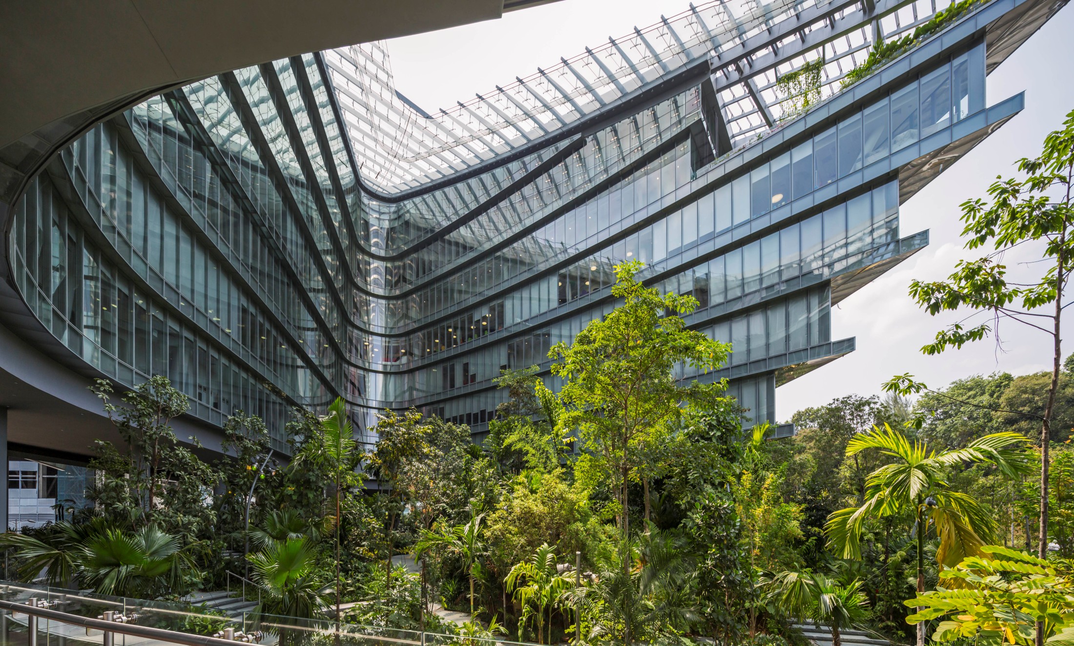 Aedas’ Eclipse (Sandcrawler) in Singapore, a V‑shaped office complex with stepped glass wings, aluminium louvres and planted courtyard. Oblique view into Aedas’ nine‑storey Eclipse complex showing two tapering office wings forming a bent V shape. The façades use floor‑to‑ceiling glazing and horizontal aluminium louvres, with stepped overhangs at each level. A glazed roof spans part of the central void, enclosing a densely planted landscape courtyard.