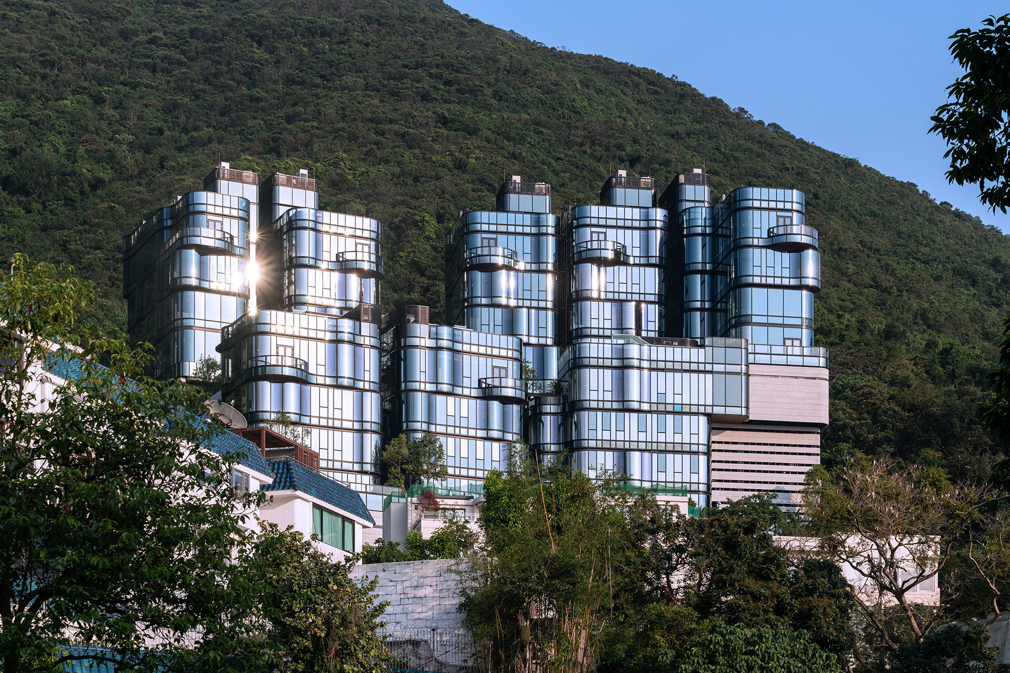 Aedas’ Pulsa residential villas with sculptural curved glass façades cascading above Repulse Bay, Hong Kong. Oblique hillside view of Aedas’ Pulsa, an eight‑house residential development composed of stepped, low‑rise villa volumes. The buildings feature floor‑to‑ceiling glass curtain walls with pronounced curved corners and horizontal slab lines. Perched against a vegetated slope, the houses are oriented toward sea views within a subtropical coastal context.