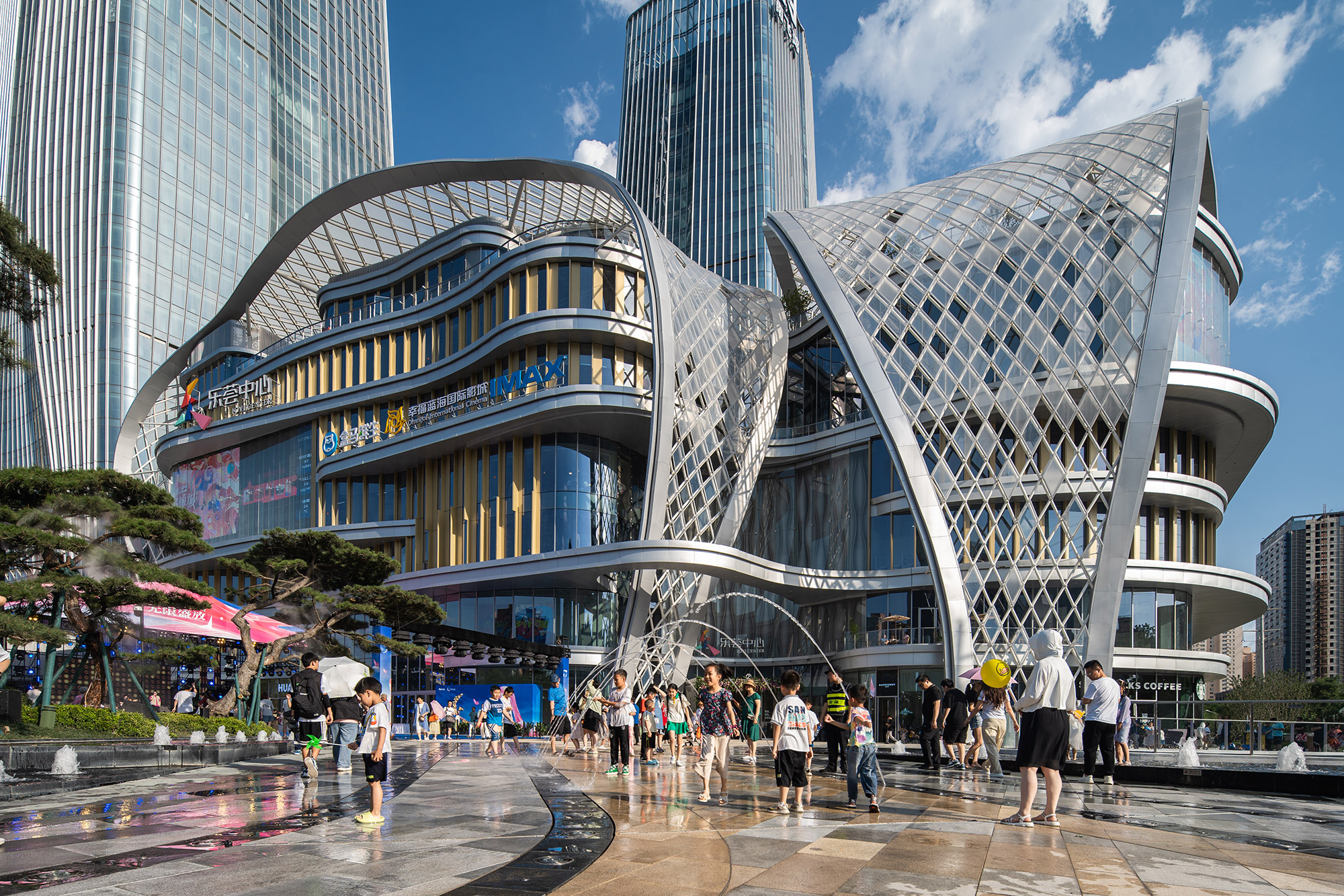 Aedas’ mixed‑use retail podium with free‑form steel gridshell canopy and curved glass façades within a dense urban plaza. Ground‑level view of an Aedas‑designed mixed‑use complex showing a multi‑storey retail podium with floor‑to‑ceiling glazing, metal fins, and a large free‑form steel gridshell canopy. Curved roof structures span over public space, integrating daylight, shading, and water features within an active urban plaza.