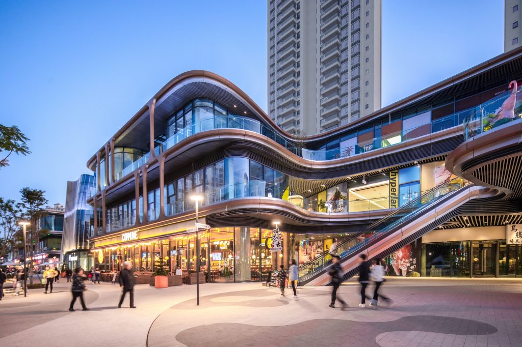 Aedas’ Chengdu Hyperlane retail podium with curving glass façades, external escalator and public plaza at dusk. Street-level view of Aedas’ Chengdu Hyperlane: a multi-storey retail podium with curvilinear floor plates, unitised glass curtain wall, deep horizontal canopy lines and a diagonal external escalator linking levels. Pedestrian plaza with lighting columns and patterned paving sits in front; residential towers rise behind.