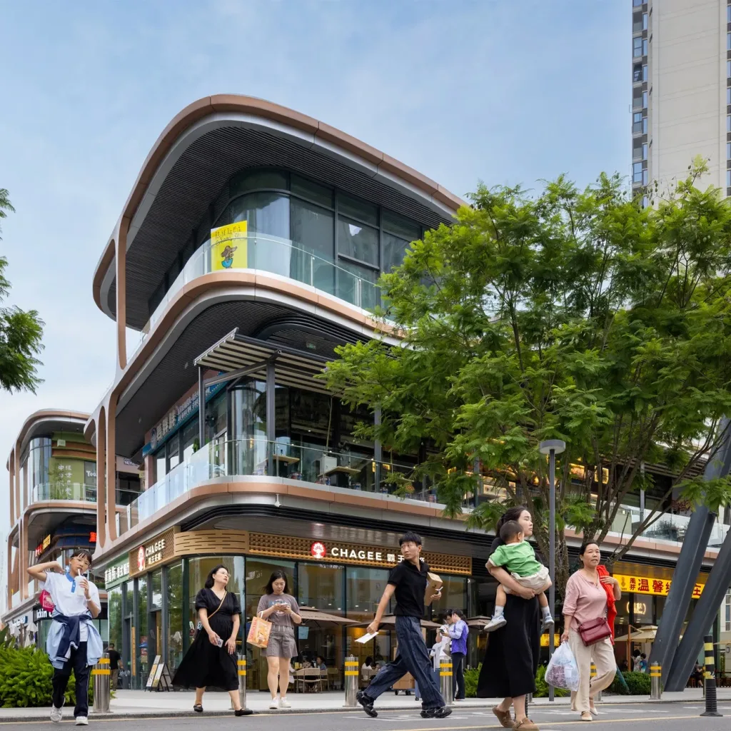 Aedas’ Chengdu Hyperlane retail podium with curving glass façades, external escalator and public plaza at dusk. Street-level view of Aedas’ Chengdu Hyperlane: a multi-storey retail podium with curvilinear floor plates, unitised glass curtain wall, deep horizontal canopy lines and a diagonal external escalator linking levels. Pedestrian plaza with lighting columns and patterned paving sits in front; residential towers rise behind.