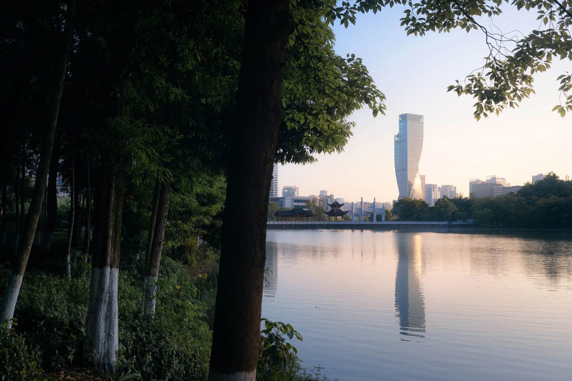 Aedas’ Chongqing Gaoke Group Office Project - waterfront skyline view with a curved glass office tower reflected in an urban lake at dusk. Aedas project image showing a waterfront urban precinct at dusk: an urban lake foreground with rippled reflections, a curved glass curtain-wall high-rise tower on the far shore, low-rise buildings behind trees, and a lit pavilion-like structure along the promenade. Vegetated lakeside edge frames the view.