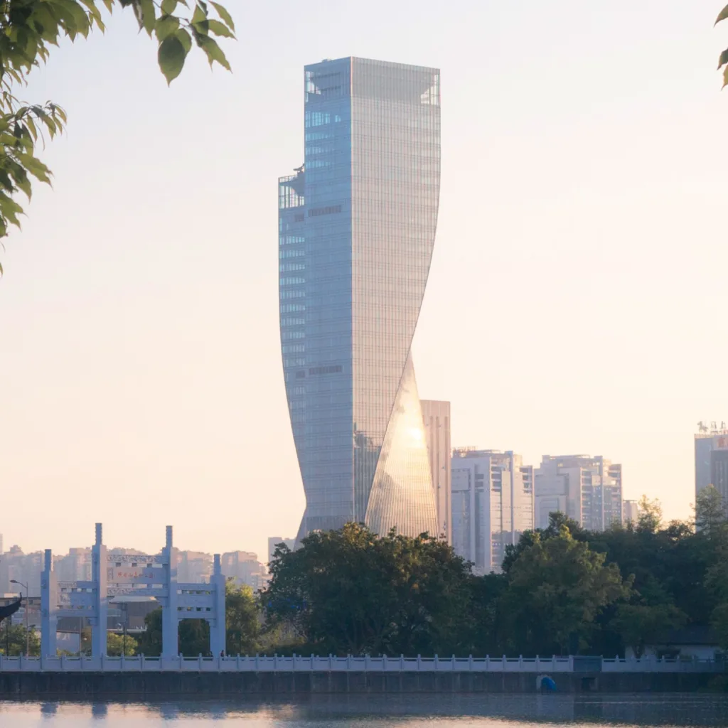 Aedas’ Chongqing Gaoke Group Office Project - waterfront skyline view with a curved glass office tower reflected in an urban lake at dusk. Aedas project image showing a waterfront urban precinct at dusk: an urban lake foreground with rippled reflections, a curved glass curtain-wall high-rise tower on the far shore, low-rise buildings behind trees, and a lit pavilion-like structure along the promenade. Vegetated lakeside edge frames the view.