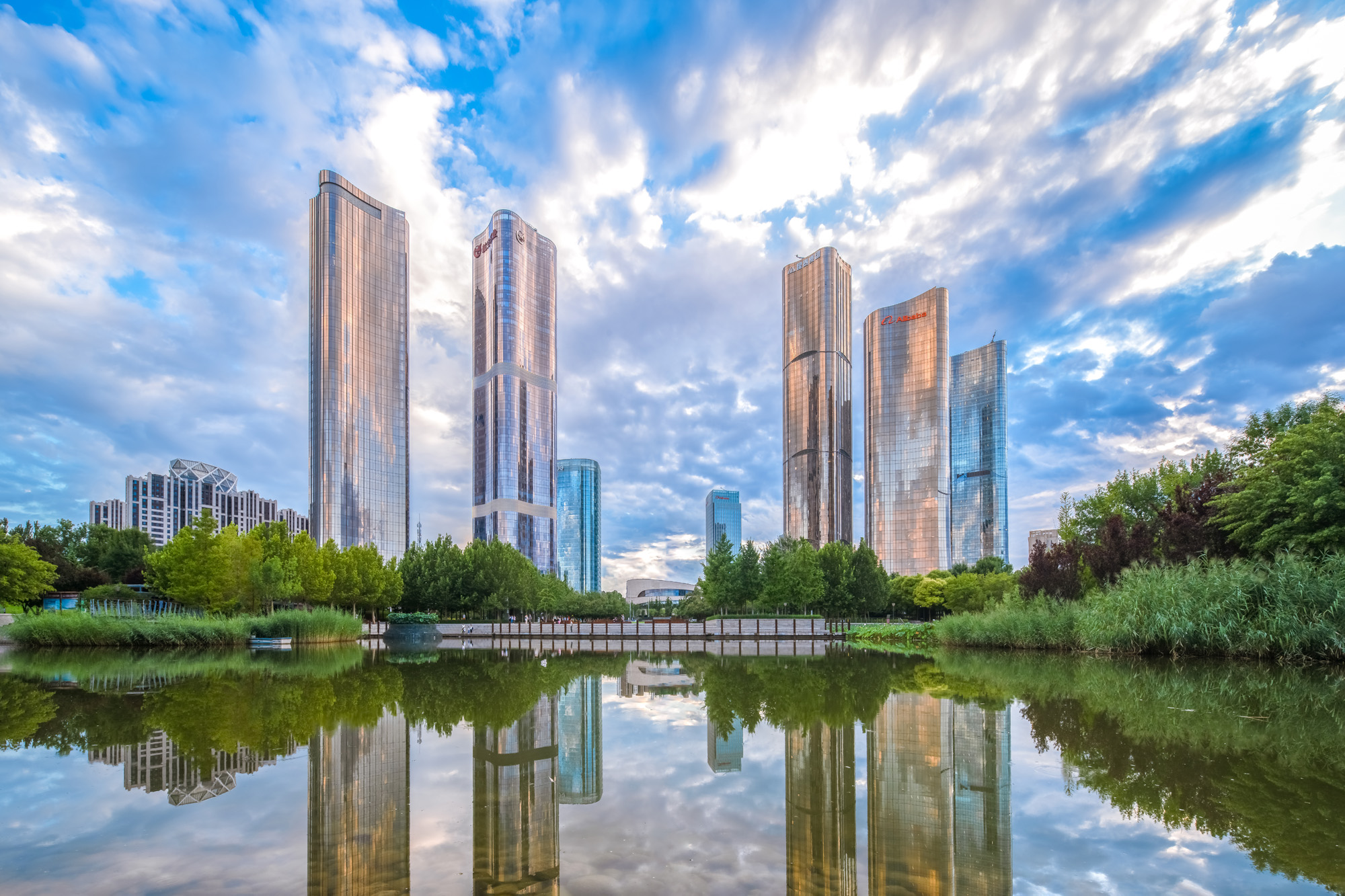 Aedas’ Da Wang Jing Plot#2 mixed-use high-rise complex with curved glazed office towers, landscaped podiums and urban green links. Aerial view of Aedas’ Da Wang Jing Plot#2 mixed-use development comprising four Grade‑A office towers and a residential tower with continuous high-performance curtain wall glazing. Curvilinear podiums integrate commercial and exhibition spaces, planted roof decks, internal roads and landscaped buffers forming a permeable urban oasis condition.