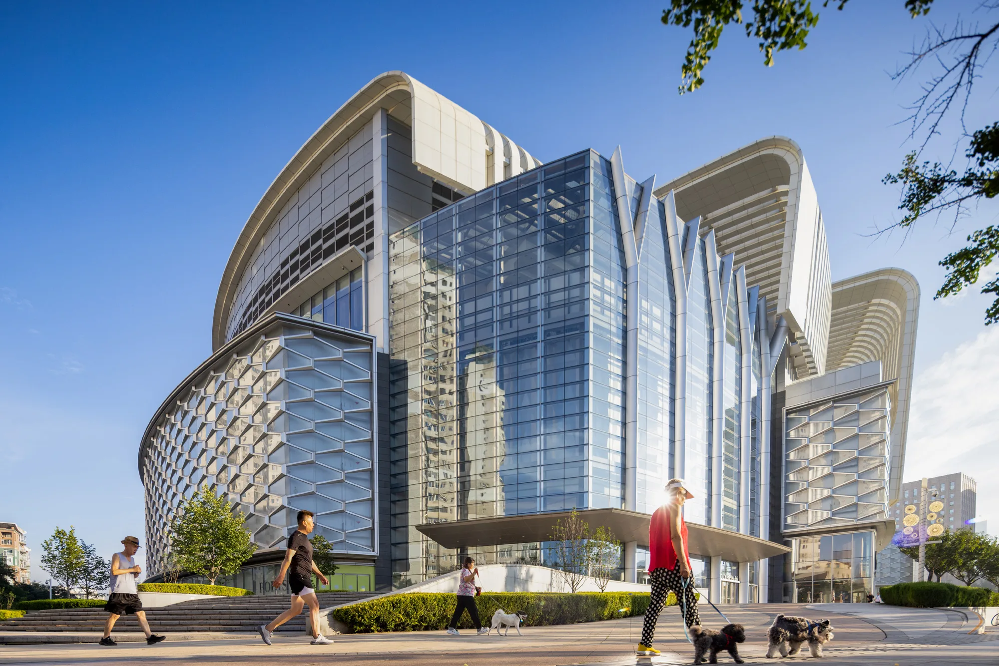 Aedas’ Olympia 66 retail complex with layered roof shells, full‑height atrium glazing and curving façade geometry. Street‑level view of Aedas’ Olympia 66 showing a large retail complex with curved roof shells stacked in layers above clear storey glazing. A central glazed atrium façade is flanked by metal‑clad volumes. The podium integrates recessed entrances, horizontal shading elements, and a pedestrian plaza, forming a landmark retail development within a dense urban environment.
