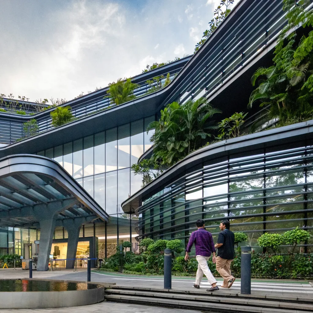 Aedas’ Unilever HQ Jakarta with curved louvred façade, glazed atrium and planted terraces in BSD Green Office Park. Exterior view of Aedas’ Unilever Headquarters in Jakarta: a low-rise office building with rounded corners, continuous horizontal sun‑shading louvres, and floor‑to‑ceiling glazing facing a covered drop‑off canopy. Planted roof terraces and perimeter landscaping frame the entry forecourt, supporting a green‑district campus setting.
