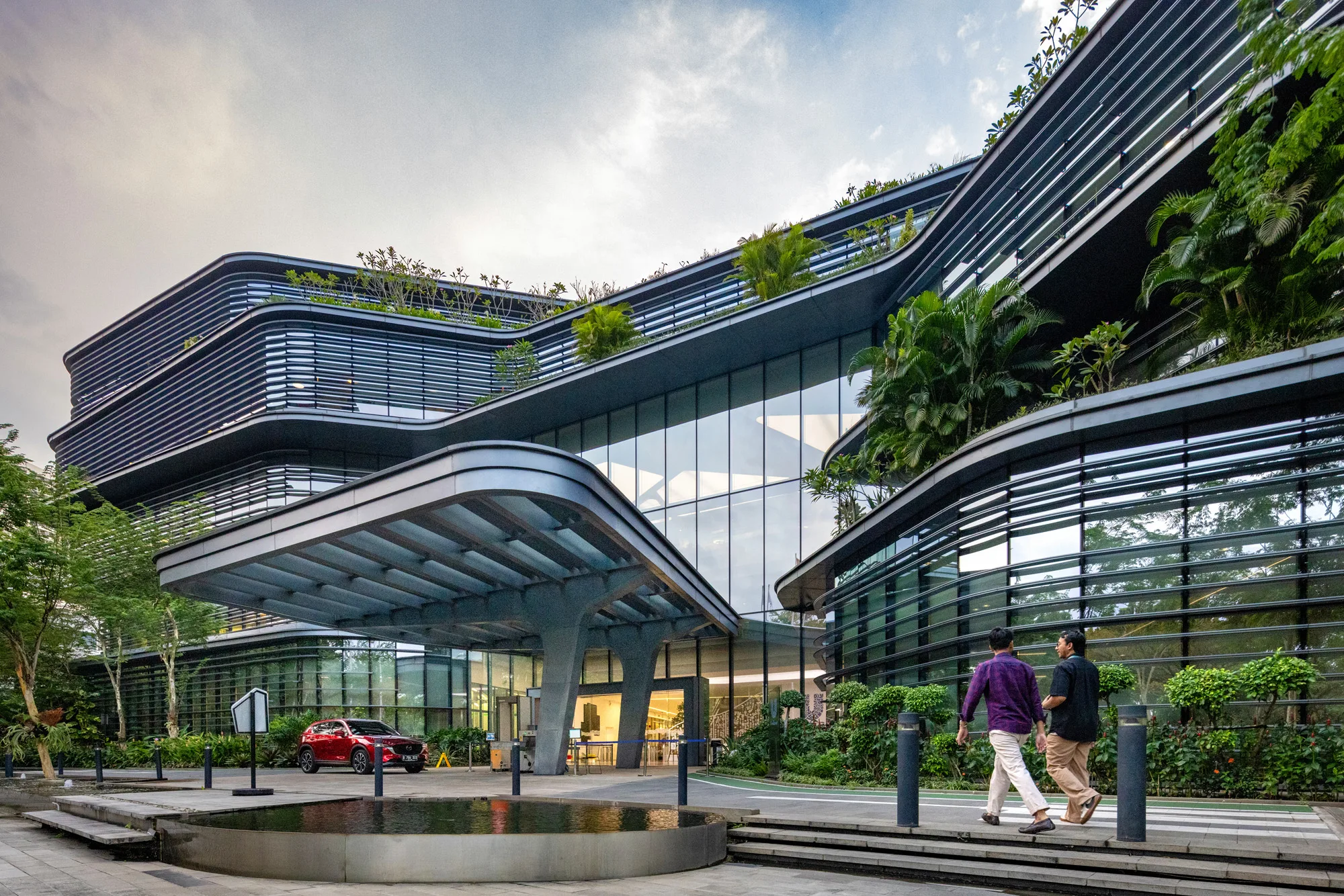 Aedas’ Unilever HQ Jakarta with curved louvred façade, glazed atrium and planted terraces in BSD Green Office Park. Exterior view of Aedas’ Unilever Headquarters in Jakarta: a low-rise office building with rounded corners, continuous horizontal sun‑shading louvres, and floor‑to‑ceiling glazing facing a covered drop‑off canopy. Planted roof terraces and perimeter landscaping frame the entry forecourt, supporting a green‑district campus setting.