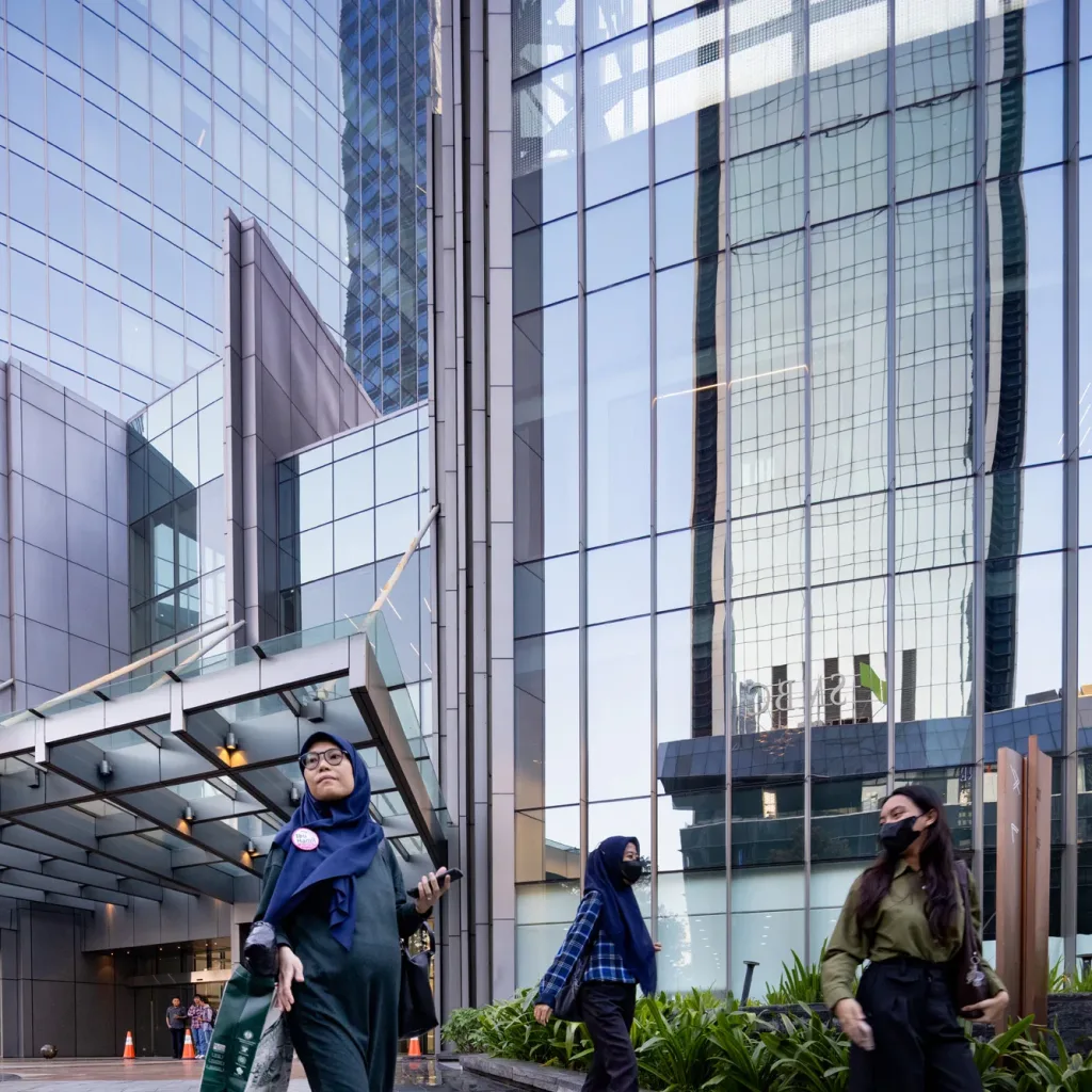 Aedas’ World Capital Tower in Jakarta with curved glass curtain wall, podium canopy and streetscape landscaping in Mega Kuningan CBD. Street‑level view of the Aedas‑designed World Capital Tower in Jakarta, showing a high‑rise commercial tower with vertically curved glass curtain wall above a podium retail base. A projecting entrance canopy, reflective façade panels and integrated planted forecourt interface with a multi‑lane CBD street, framed by surrounding high‑density urban development.