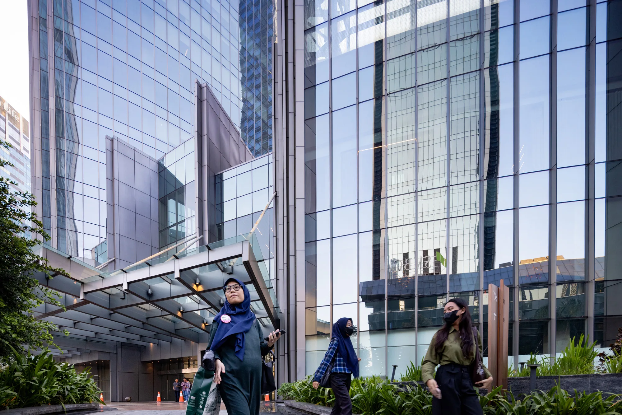 Aedas’ World Capital Tower in Jakarta with curved glass curtain wall, podium canopy and streetscape landscaping in Mega Kuningan CBD. Street‑level view of the Aedas‑designed World Capital Tower in Jakarta, showing a high‑rise commercial tower with vertically curved glass curtain wall above a podium retail base. A projecting entrance canopy, reflective façade panels and integrated planted forecourt interface with a multi‑lane CBD street, framed by surrounding high‑density urban development.