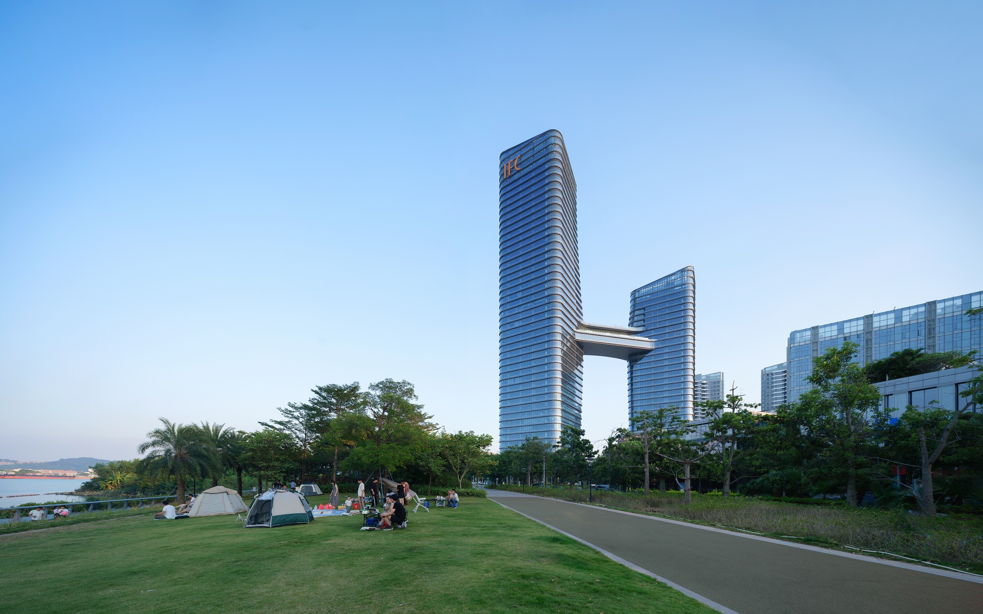 Aedas’ Nansha IFC twin office towers with slanted glass façades and sky garden overlooking Lingshan Island, Guangzhou. Long‑range view of Aedas’ Nansha IFC showing two high‑rise office towers connected by a sky garden. The towers feature unitised curtain walls with slanted glass panels and rounded corners. Set within a landscaped waterfront park, the development integrates pedestrian routes, green buffers and mixed‑use podium elements in a coastal urban environment.