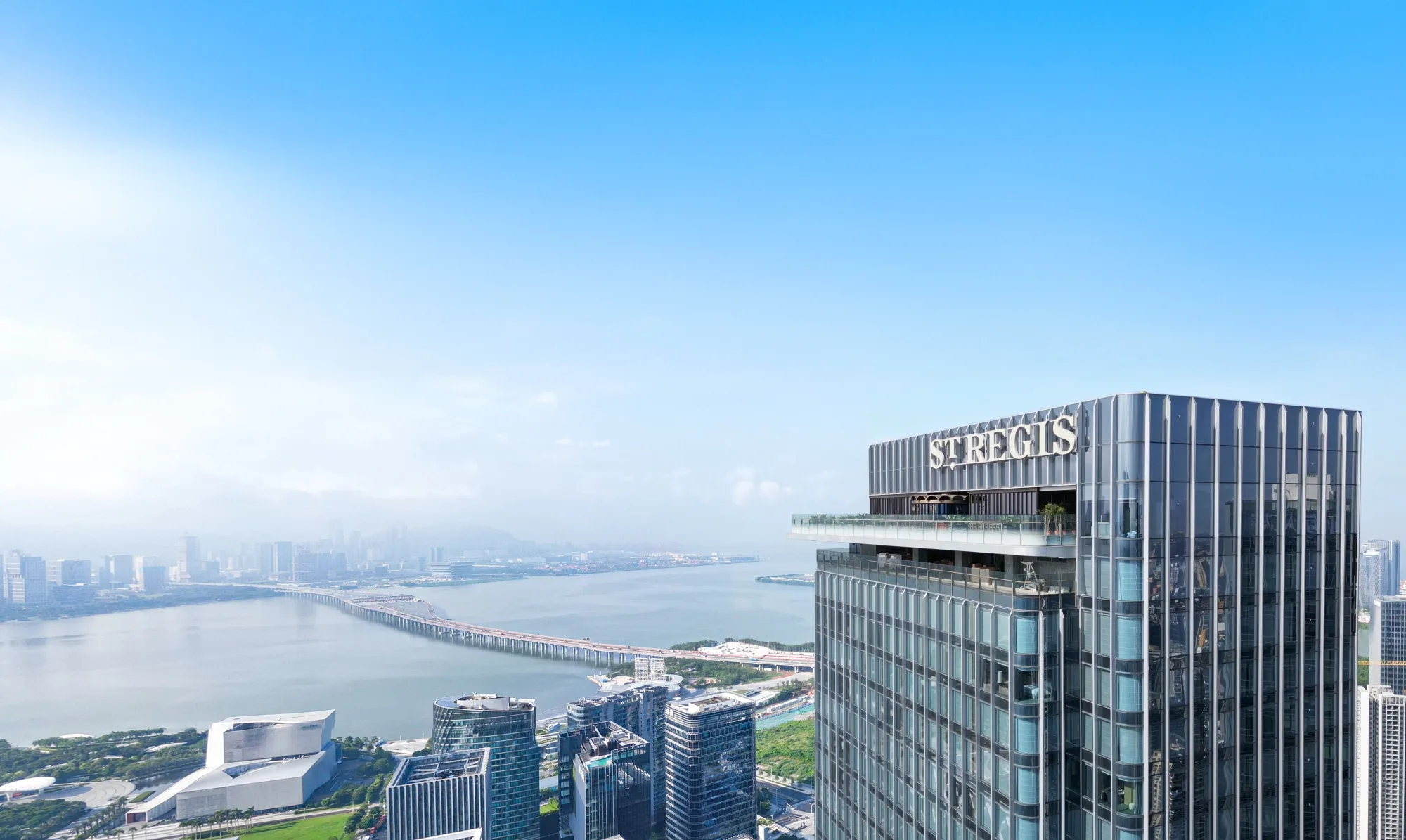 Aedas’ OCT Real One / Ruiwan Tower with vertical ribbed aluminium façade and glazed curtain wall overlooking Qianhai Bay in Shenzhen. High-rise tower by Aedas featuring a slender rectilinear form clad with glazed curtain wall systems and continuous vertical aluminium ribbing. The tower rises from a mixed-use podium with recessed glazing, set within a coastal urban district overlooking Qianhai Bay. Surrounding infrastructure and greenery indicate integration with a waterfront and central green axis.