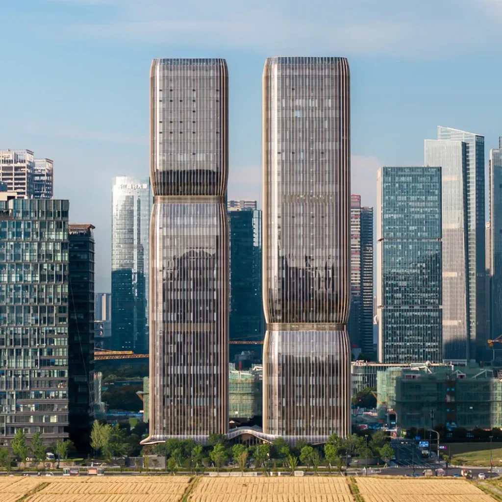 Aedas’ Hangzhou One Future twin towers with stepped, porous façades and bronze aluminium‑glass envelope in a CBD setting. Street‑level view of Aedas’ Hangzhou One Future showing two symmetrical high‑rise towers with staggered refuge floors forming stepped façades. The buildings are clad in aluminium and glass panels, producing a bronze‑toned curtain wall. A curved entrance canopy addresses the street, integrating office and residential uses within a dense urban context near metro lines.