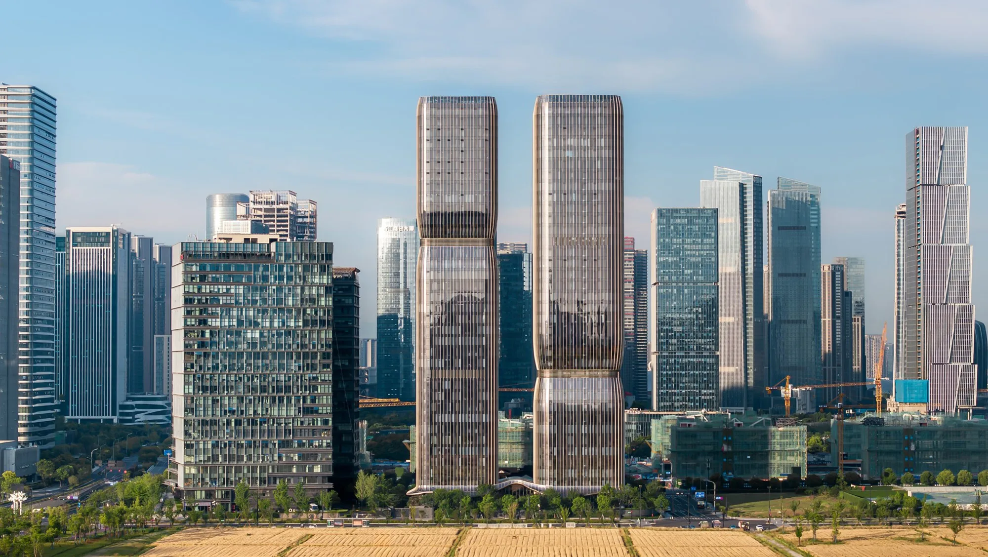 Aedas’ Hangzhou One Future twin towers with stepped, porous façades and bronze aluminium‑glass envelope in a CBD setting. Street‑level view of Aedas’ Hangzhou One Future showing two symmetrical high‑rise towers with staggered refuge floors forming stepped façades. The buildings are clad in aluminium and glass panels, producing a bronze‑toned curtain wall. A curved entrance canopy addresses the street, integrating office and residential uses within a dense urban context near metro lines.