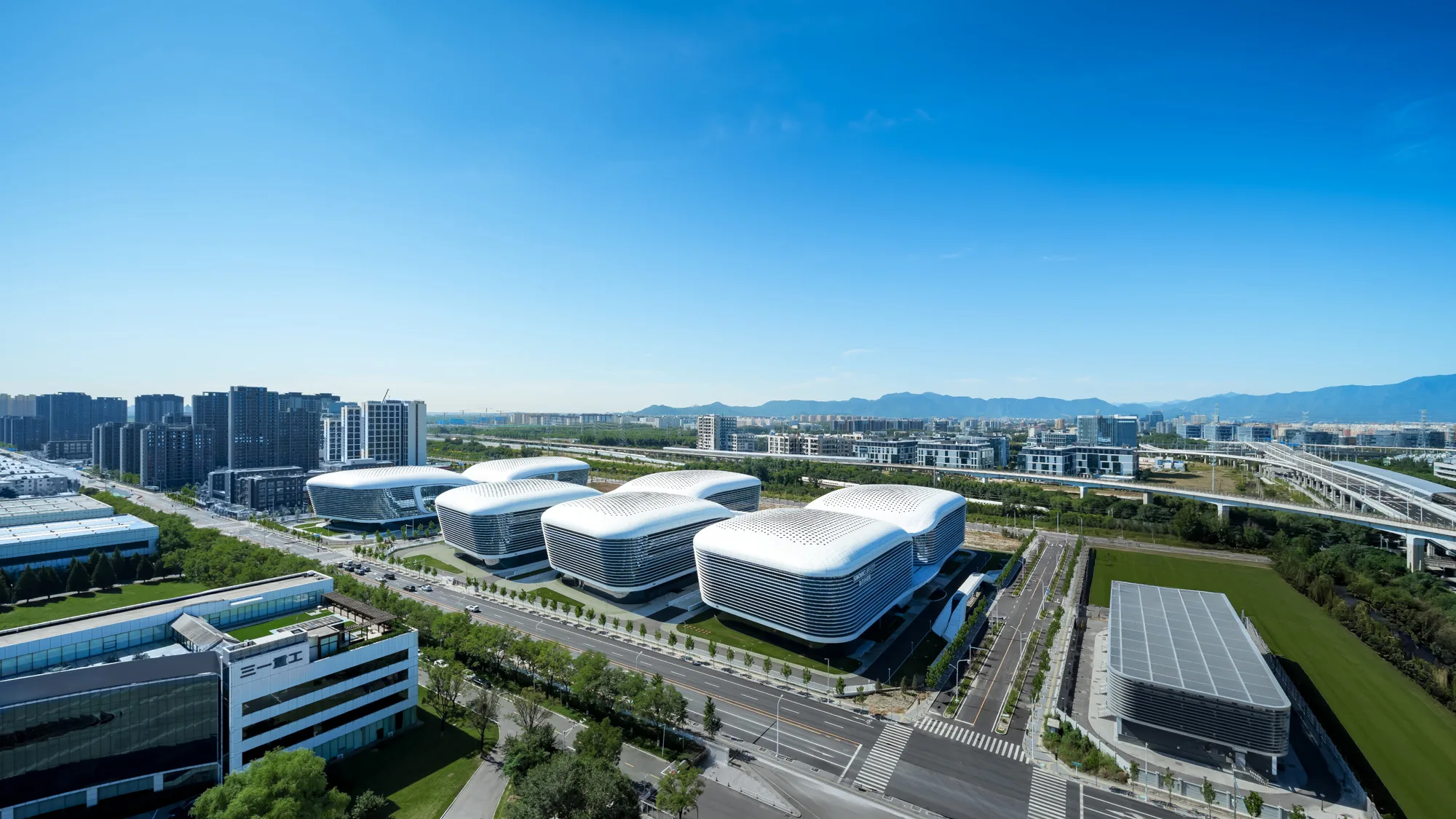 Aedas’ SINOVAC high‑tech campus with clustered R&D and manufacturing buildings linked by elevated corridors. Aerial view of an Aedas‑designed science and technology campus comprising multiple low‑rise, rounded‑corner building volumes with continuous horizontal glazing bands. The buildings are arranged in parallel rows and linked by elevated corridors above landscaped grounds. Wide internal roads, green buffers and adjacent metro and expressway infrastructure frame the campus within a suburban innovation district.
