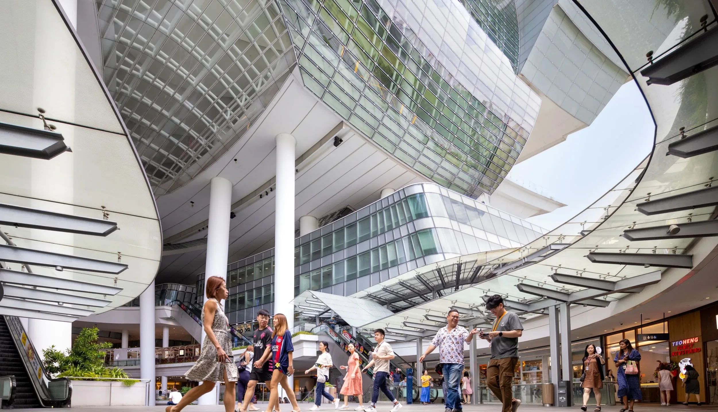 Aedas’ The Star, Singapore—covered open-air retail atrium with curved glazed canopies and pedestrian concourse. Ground-level view inside Aedas’ The Star mixed-use retail/cultural complex in Singapore: open-air mall concourse beneath curved glass-and-steel canopies, deep overhangs and tall white columns. Overhead, a suspended glazed volume and curtain-wall façade frame the public plaza. Pedestrians circulate between shopfronts—visual evidence for daylighting strategy, solar control and tropical passive design.
