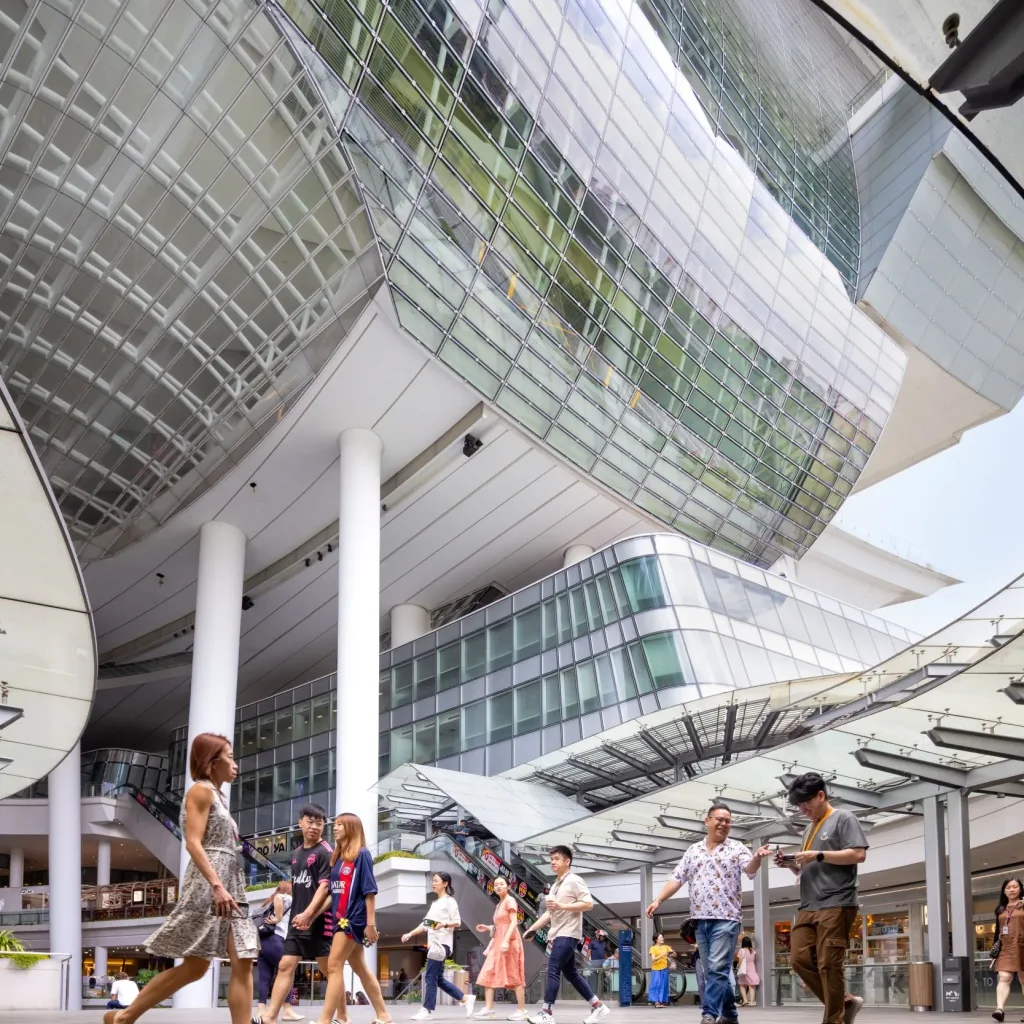 Aedas’ The Star, Singapore—covered open-air retail atrium with curved glazed canopies and pedestrian concourse. Ground-level view inside Aedas’ The Star mixed-use retail/cultural complex in Singapore: open-air mall concourse beneath curved glass-and-steel canopies, deep overhangs and tall white columns. Overhead, a suspended glazed volume and curtain-wall façade frame the public plaza. Pedestrians circulate between shopfronts—visual evidence for daylighting strategy, solar control and tropical passive design.