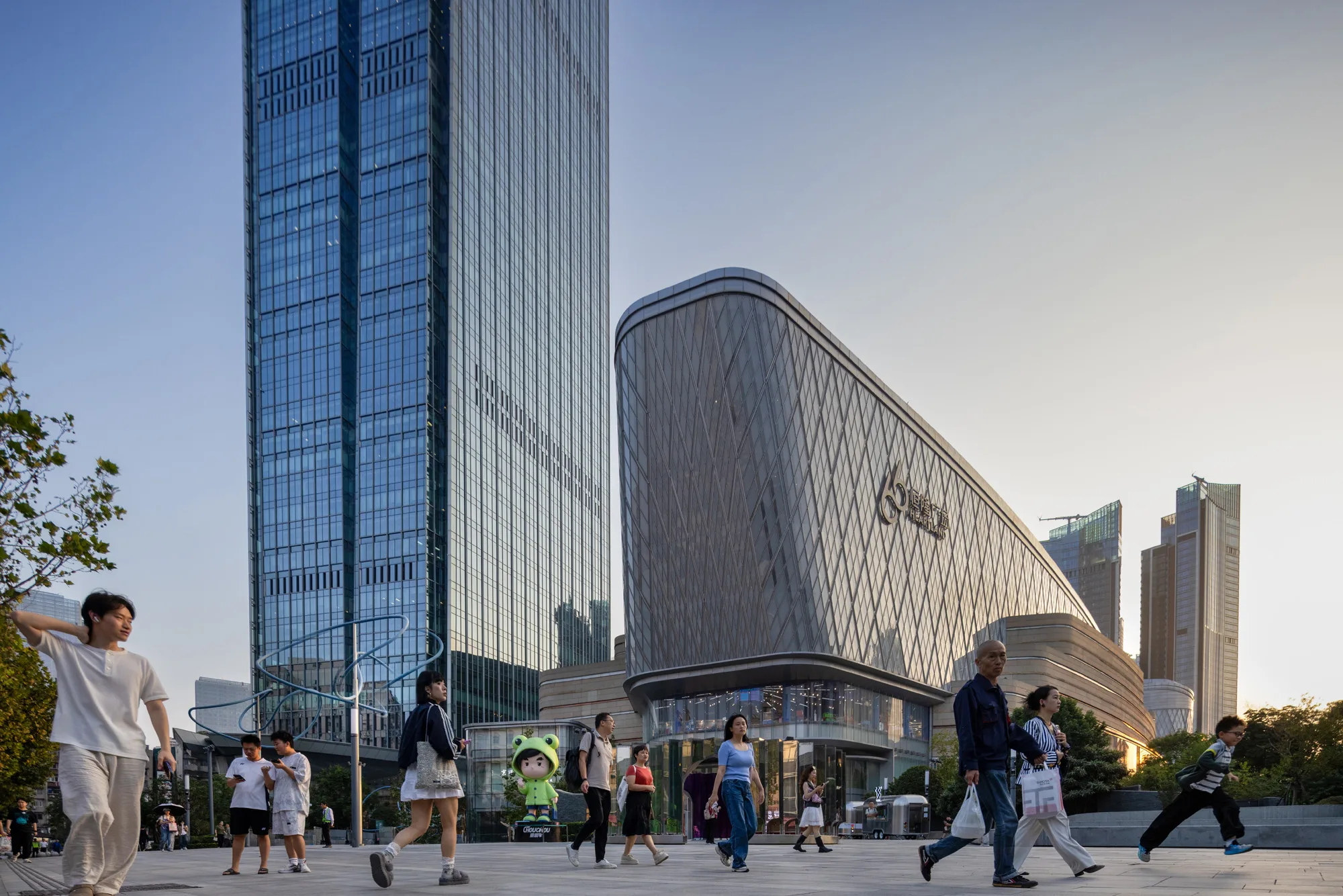 Aedas’ Heartland 66 mixed‑use development with curved retail podium and high‑rise towers in Wuhan’s CBD. Street‑level view of Aedas’ Heartland 66 showing a curvilinear retail podium with a diamond‑pattern metal façade beneath adjacent high‑rise towers. The podium integrates recessed glazing at ground level and stepped terraces above. Pedestrian hardscape and public forecourt activate the streetscape within Wuhan’s dense, transit‑served urban environment.