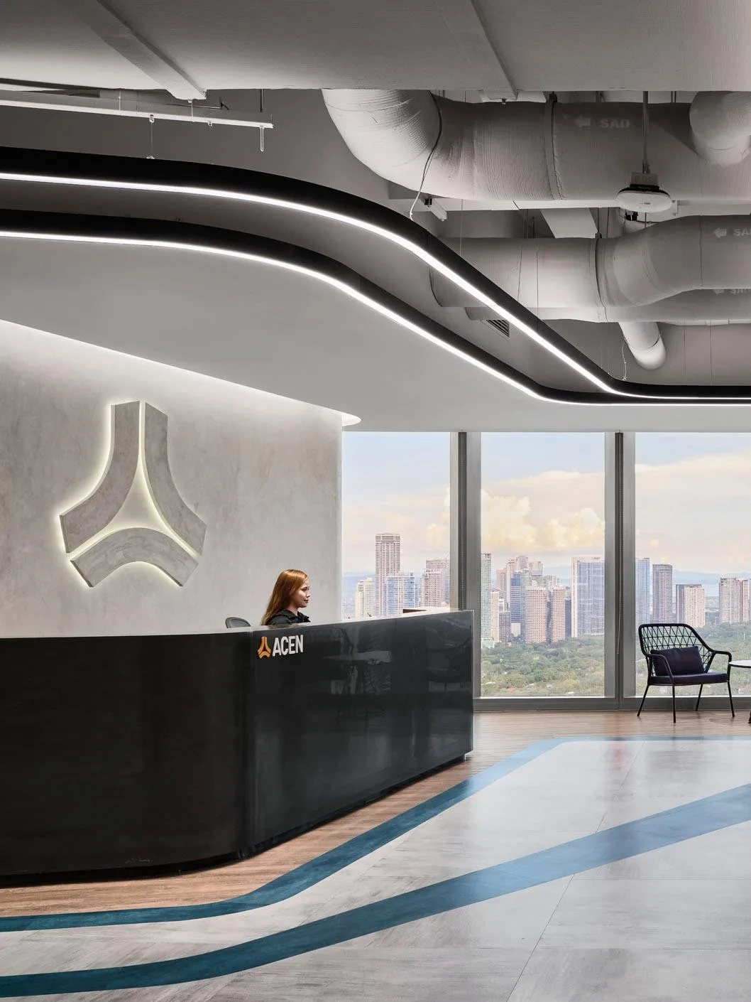 Aedas’s ACEN office in Manila is shown with a contemporary reception and social interior featuring a curved reception counter, vertically layered timber wall, fluid LED ceiling lighting, exposed services, and floor graphics suggesting energy flow. The image highlights sustainability‑driven corporate interior architecture and adaptable workplace design.
