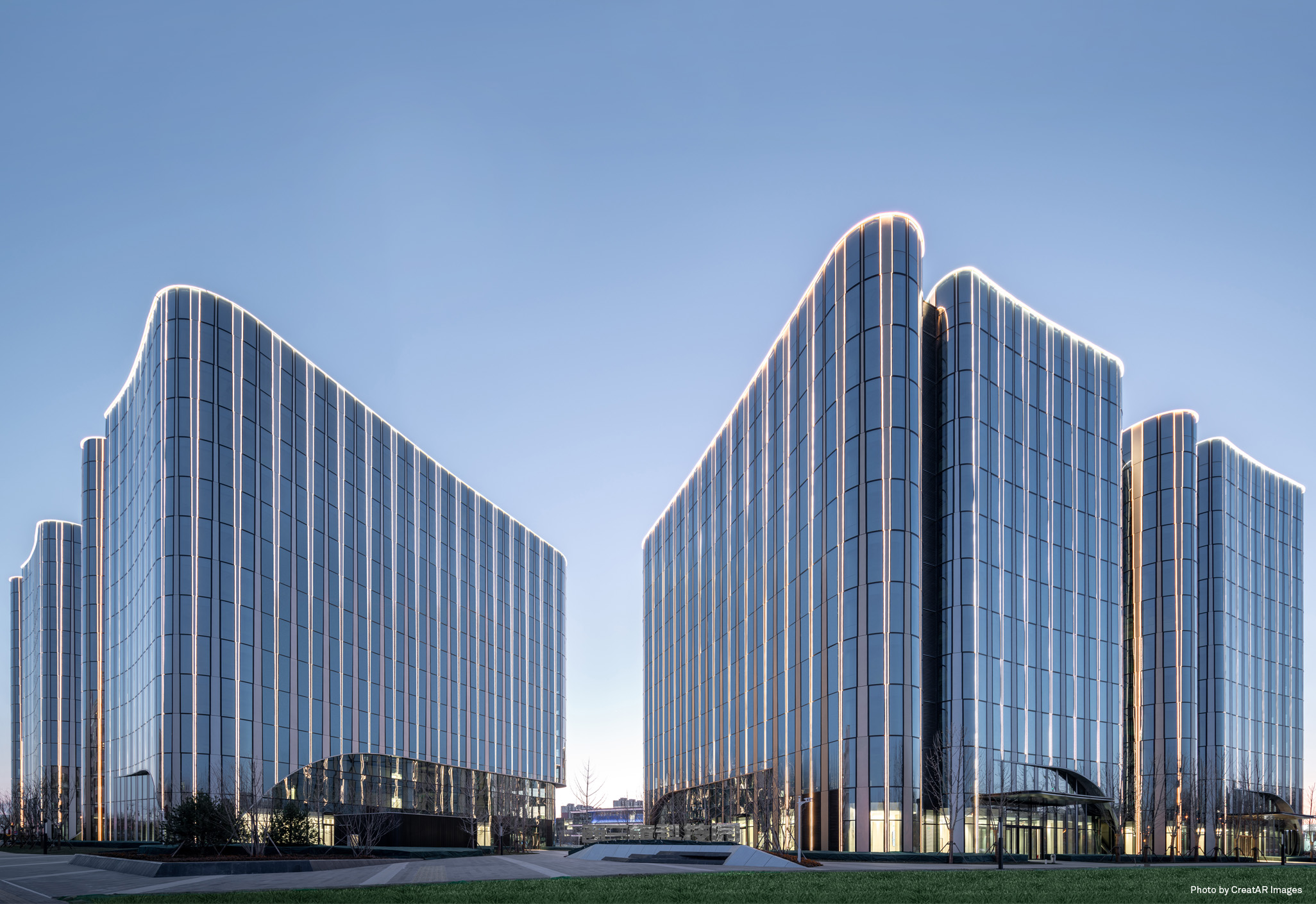Aedas’ BBMG Xi San Qi Technology Park with curved glass office blocks enclosing a central landscaped green promenade. Ground‑level dusk view of the Aedas‑designed Xi San Qi Science & Technology Park in Haidian, Beijing. The image shows multiple mid‑rise office and R&D buildings with curved floor‑to‑ceiling glass façades arranged around a central green park. Continuous vertical mullions, rounded corners and transparent podium lobbies define a pedestrian‑oriented technology campus within an urban high‑tech district.