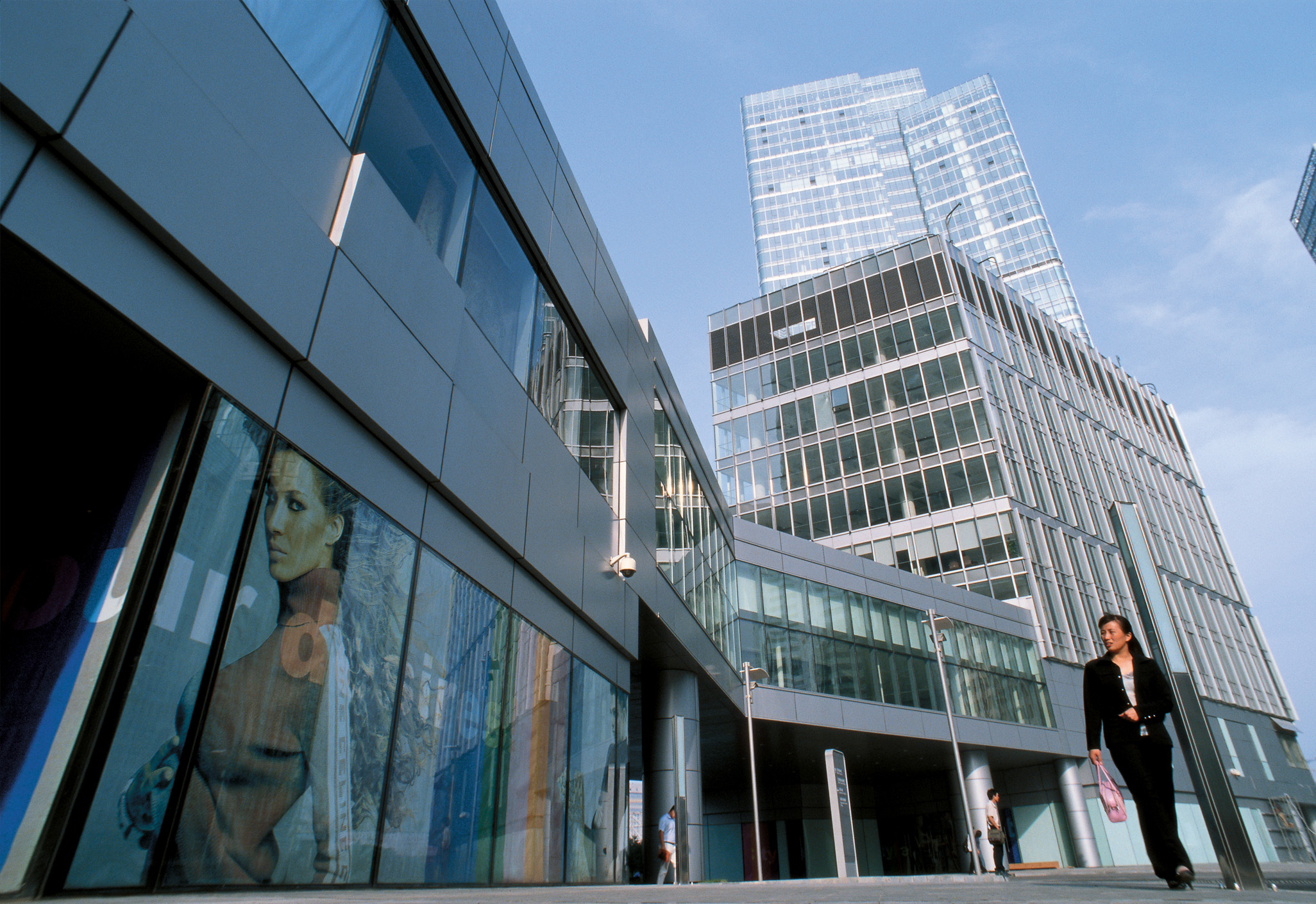 Aedas’s Fortune Plaza Phase 1 in Beijing is shown as a mixed‑use CBD development featuring stepped glass office and residential towers rising from a low‑rise retail podium. The street‑level view highlights transparent curtain walls, articulated massing, and active pedestrian frontage within Beijing’s central business district.