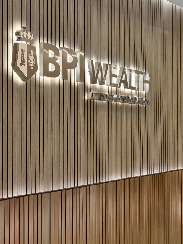 Aedas’s Bank of The Philippine Islands interior in Manila is shown with a sculpted reception area featuring vertically ribbed timber wall panels, a layered curved counter, and integrated illuminated BPI Wealth signage. The image highlights refined material detailing, corporate brand expression, acoustic wall treatment, and a contemporary banking workplace interior.