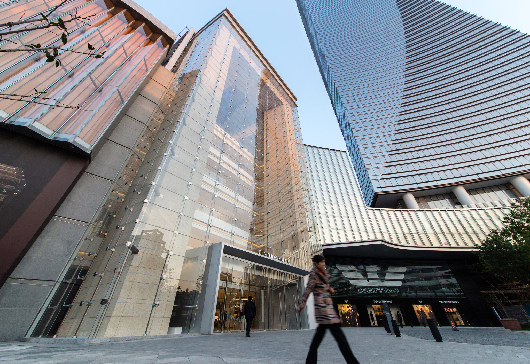 Aedas’s Center 66 Phase 1 in Wuxi is shown as a mixed‑use development with a large glazed retail atrium, horizontally layered mall podium, and a slender high‑rise office tower. The street‑level view highlights transparent façades, active retail frontage, and integration of public space within the city centre.