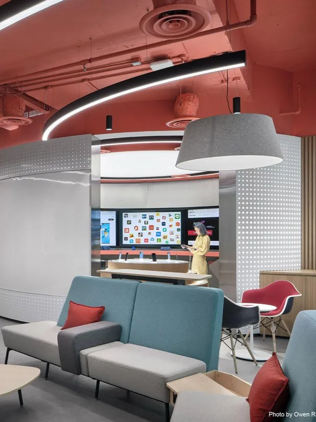 Aedas Interiors’ DIGIX Lab in Singapore is shown as an experiential technology showroom with a bold coral‑painted exposed ceiling, curved LED lighting, modular seating, and interactive app display zones. The image highlights flexible interior planning, strong brand colour integration, and a hybrid environment supporting collaboration, demonstration, and training functions.