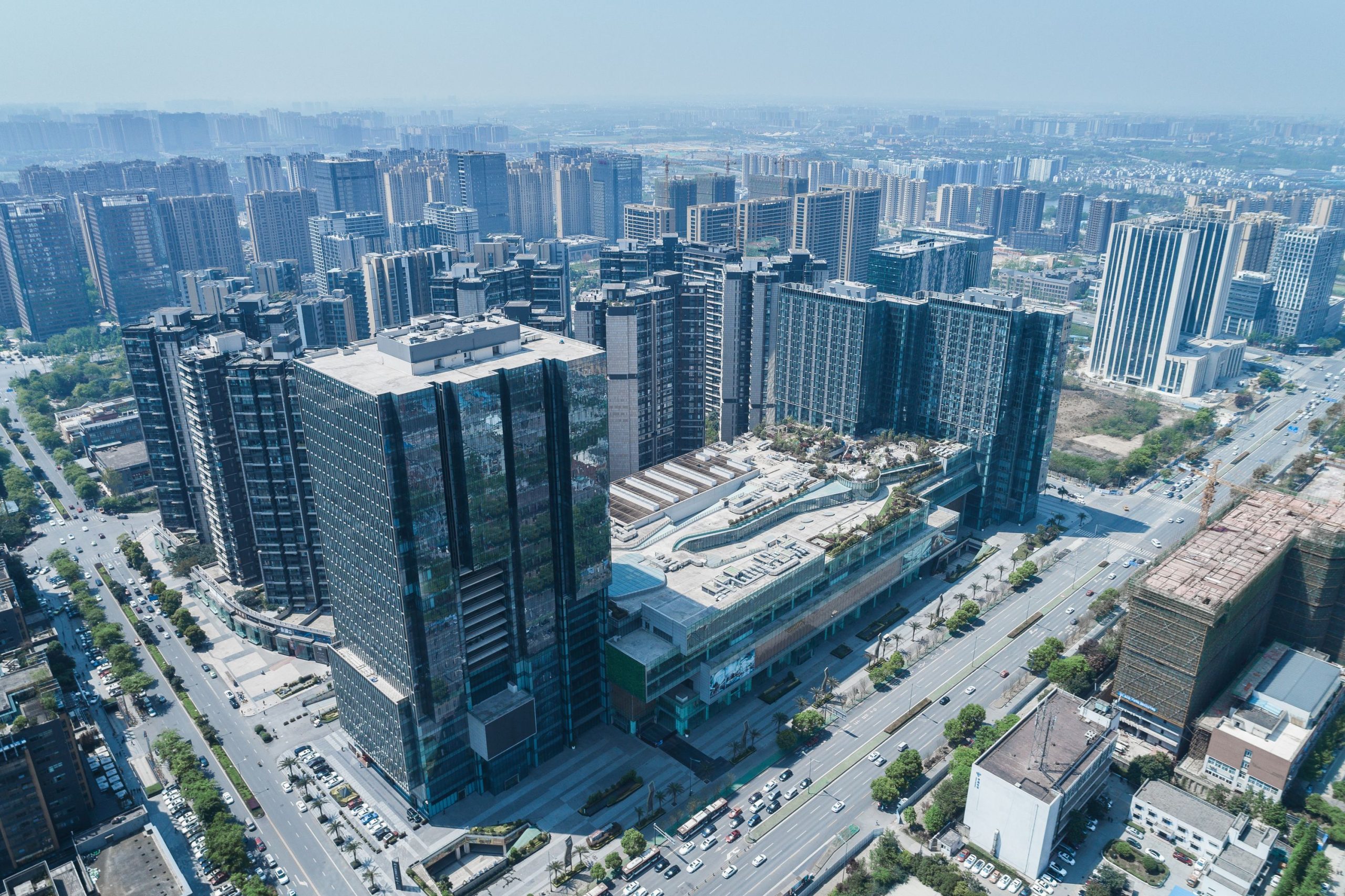 Aedas’ Chengdu mixed‑use development with layered office tower, serviced apartments and a terraced retail podium. Aerial view of an Aedas‑designed mixed‑use complex in Chengdu, showing a tall office tower with staggered, slice‑shaped floor plates rising from a multi‑storey retail podium. Serviced apartment towers are positioned along the site edge. Extensive glass curtain walls, stepped massing, and rooftop terraces frame major urban roads and surrounding residential districts.
