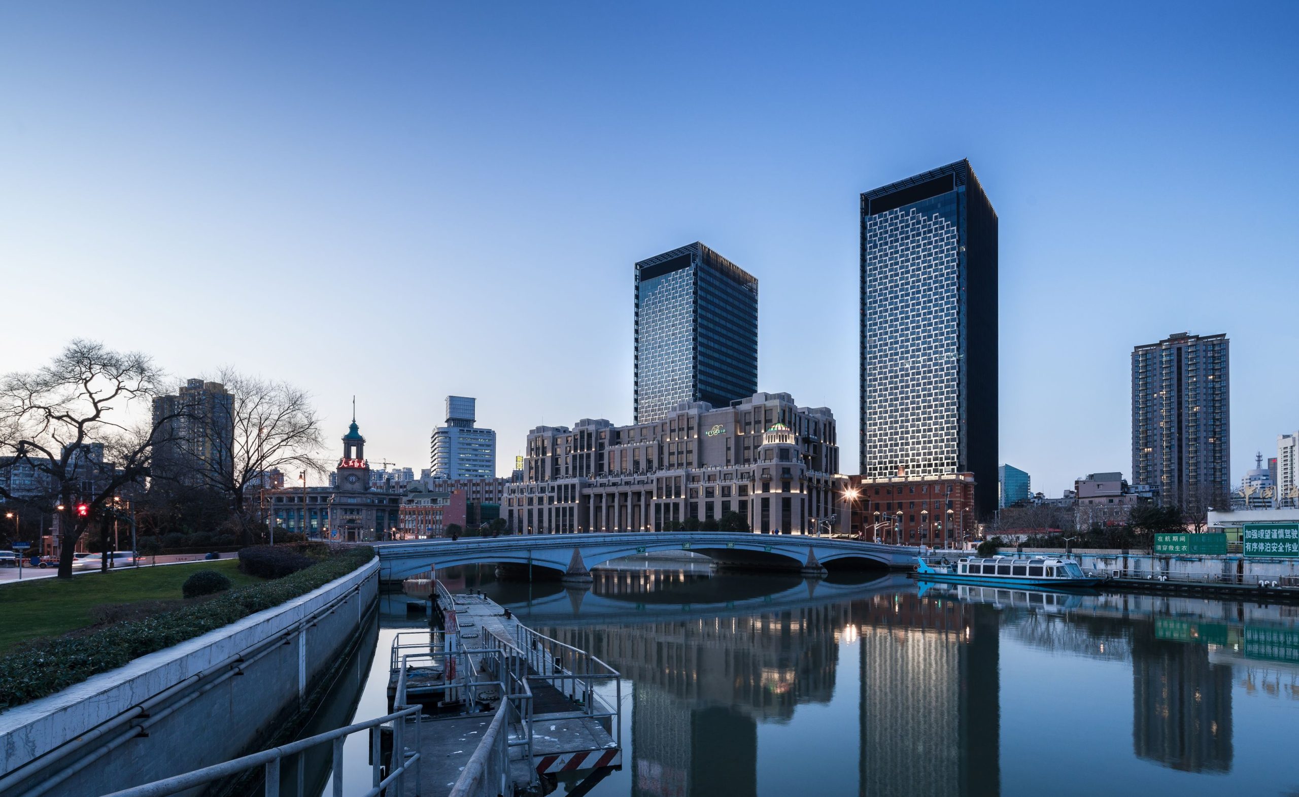 Aedas’ Shanghai Landmark Center twin office towers with patterned glass façades overlooking Suzhou Creek. Riverfront view of the Aedas‑designed Shanghai Landmark Center, showing two slender rectangular office towers rising from a low‑rise retail base. The façades feature a repetitive, lattice‑like pattern integrated with floor‑to‑ceiling glazing. The development sits along Suzhou Creek, framed by historic structures, bridges and pedestrian waterfront infrastructure.