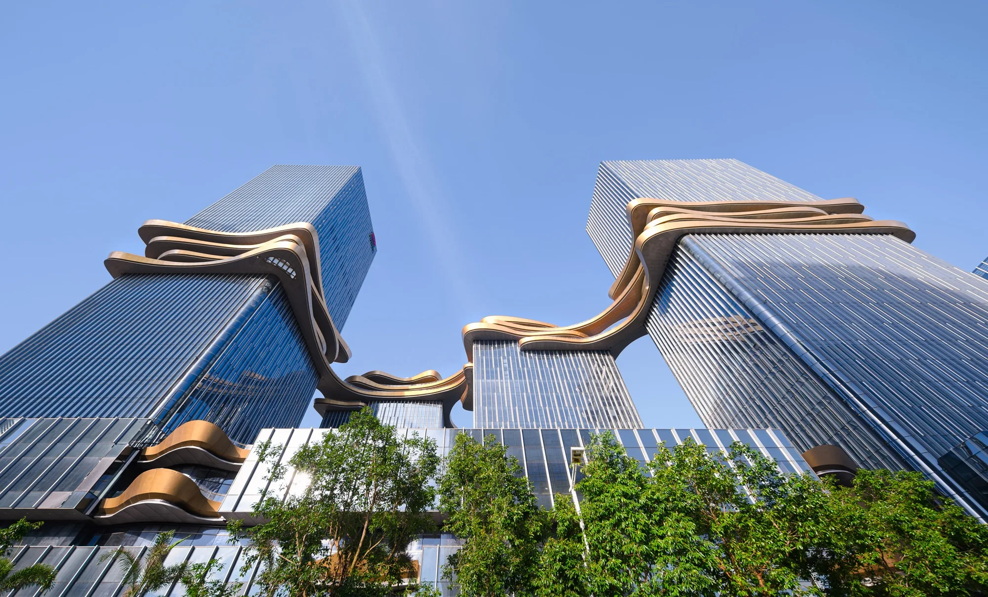 Aedas’ Hengqin CRCC Plaza in Zhuhai is shown as a dynamic office complex formed by four crystalline towers linked by a sculptural sky bridge. The image highlights vertically ribbed glass façades wrapped with flowing metallic bands, creating a continuous “loop in the sky.” Integrated greenery, roof gardens, and a layered podium activate the public realm, establishing a distinctive business landmark within the emerging central business district.