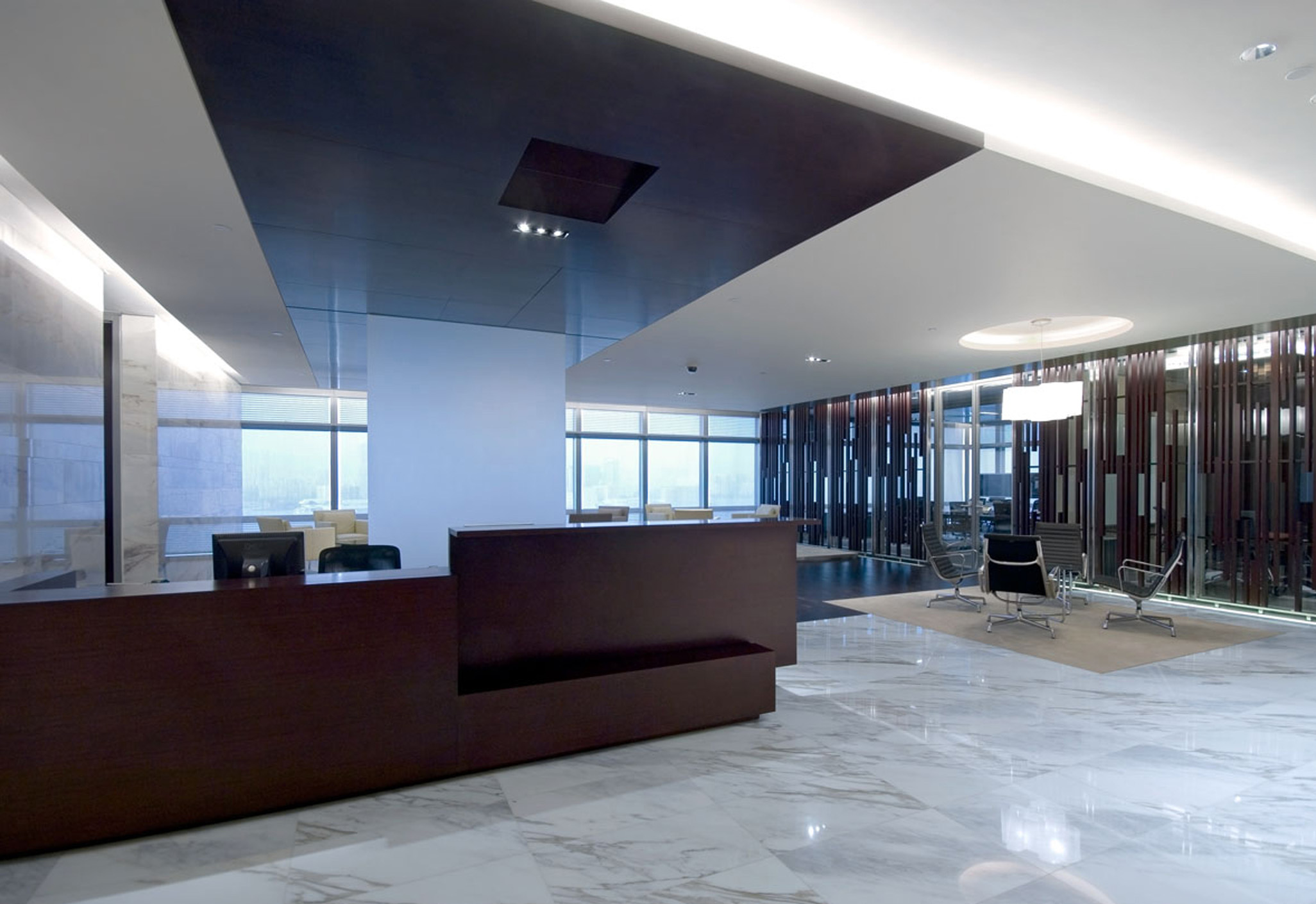 Aedas Interiors’ AIA Asia Headquarters in Hong Kong is shown with an executive reception and lounge environment featuring a dark timber reception counter, polished marble flooring, and semi‑transparent timber screen walls. The image highlights refined corporate interior architecture that blends neutral tones with Asian material influences, creating a calm, prestigious workspace with visual openness and hierarchical zoning across interconnected executive floors.