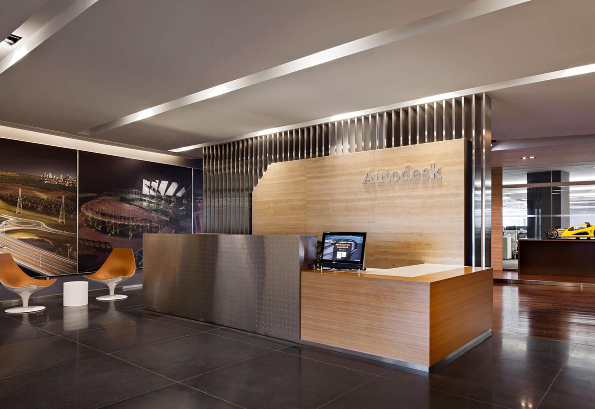 Aedas Interiors’ Autodesk Asia office in Singapore is shown with a refined reception area featuring a timber‑clad counter, perforated metal screen, integrated branding wall, and dark stone flooring. The image highlights a neighbourhood‑inspired workplace interior, combining clean architectural lines, layered materials, and open visual connectivity to support collaboration, client engagement, and a contemporary regional headquarters environment.