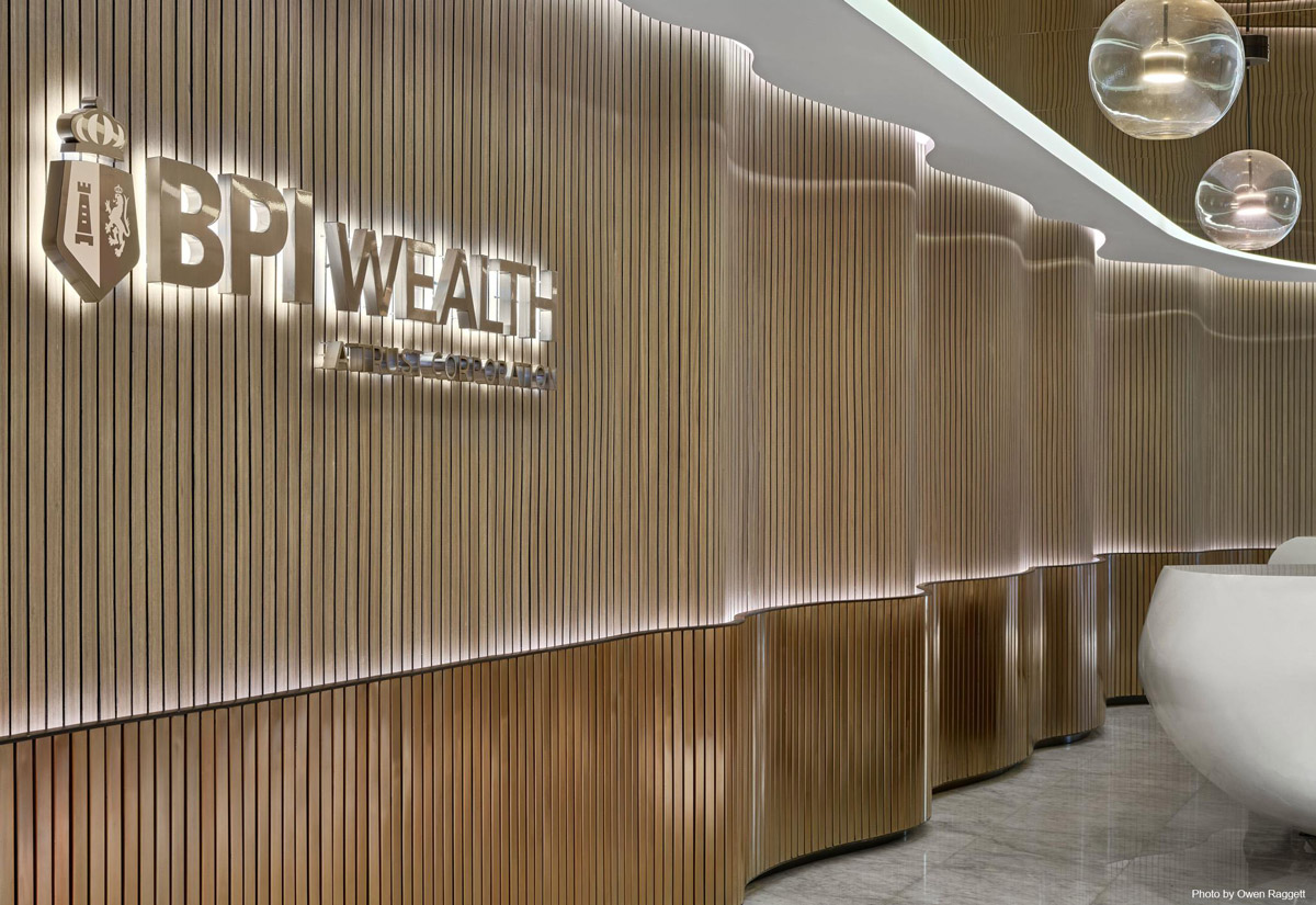 Aedas’s Bank of The Philippine Islands interior in Manila is shown with a sculpted reception area featuring vertically ribbed timber wall panels, a layered curved counter, and integrated illuminated BPI Wealth signage. The image highlights refined material detailing, corporate brand expression, acoustic wall treatment, and a contemporary banking workplace interior.
