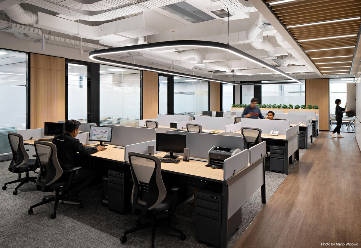 Aedas’s Napan Persada office in Jakarta is shown as a contemporary open‑plan workplace with customised workstations, integrated timber feature walls, exposed services ceiling, and linear suspended lighting. The image highlights client‑fabricated furniture, modular desk systems, and a balanced layout supporting collaboration and focused work within a CBD office environment.