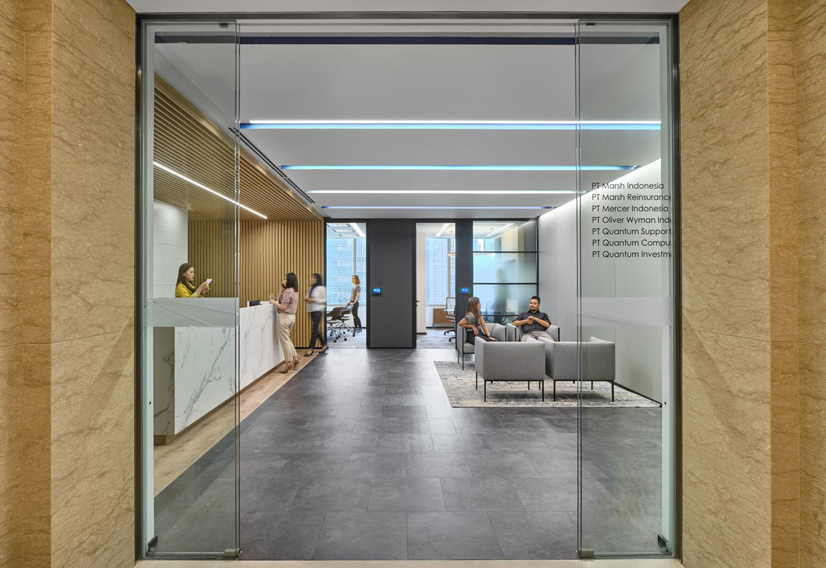 Aedas Interiors’ Marsh & McLennan Jakarta Smart Office is shown as a contemporary, glass‑fronted staff hub with integrated reception, lounge seating, and open circulation. The image highlights an agile, unassigned workplace interior combining neutral tones, timber accents, and local materials, designed to support connectivity, informal collaboration, and diverse work settings within a vibrant, technology‑enabled corporate environment.