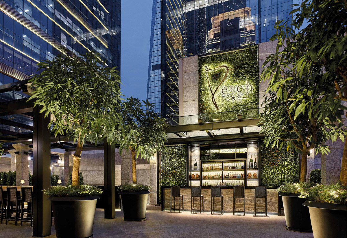 Aedas Interiors’ Perch at The Langham Shenzhen is shown as an elevated outdoor bar with lush potted trees, a green living wall façade, and a central illuminated bar pavilion. The image highlights a refined hospitality environment that blends landscape elements, warm lighting, and contemporary stone detailing, creating an intimate urban terrace setting framed by surrounding high‑rise towers.