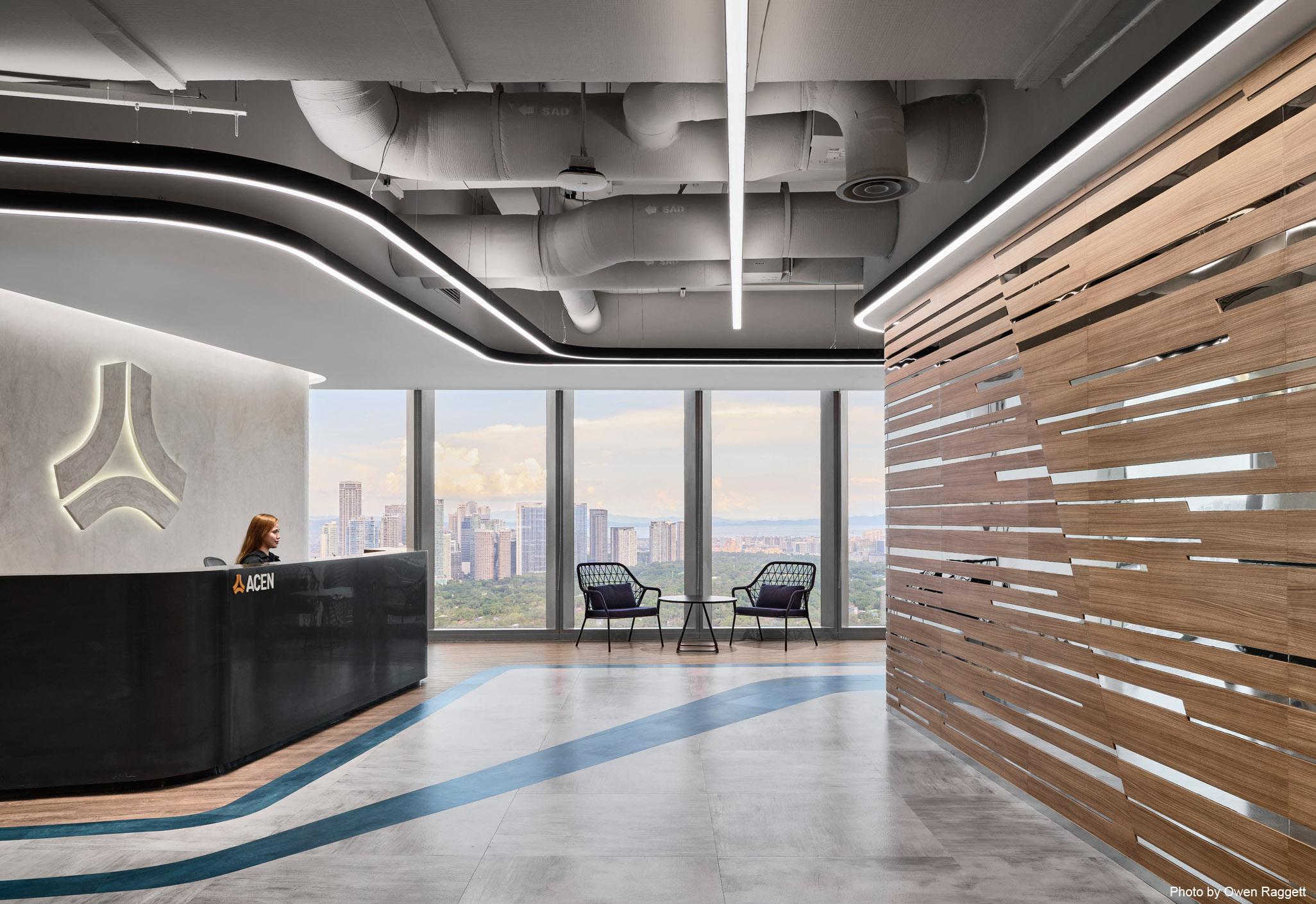 Aedas’s ACEN office in Manila is shown with a contemporary reception and social interior featuring a curved reception counter, vertically layered timber wall, fluid LED ceiling lighting, exposed services, and floor graphics suggesting energy flow. The image highlights sustainability‑driven corporate interior architecture and adaptable workplace design.
