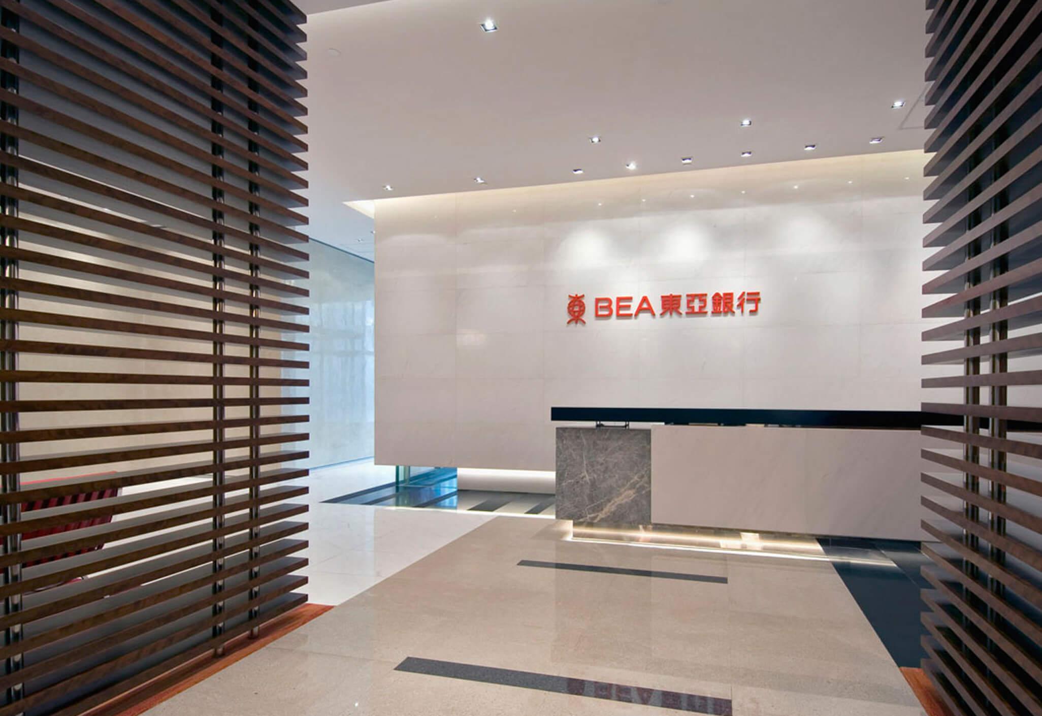 Aedas Interiors’ BEA Centralisation Project in Hong Kong is shown with a formal reception space featuring a marble‑clad desk, polished stone flooring, integrated BEA branding wall, and vertical timber screen partitions. The image highlights a refined financial‑institution interior emphasising clarity, security, and spatial organisation, supporting large‑scale departmental consolidation within a contemporary corporate headquarters environment.