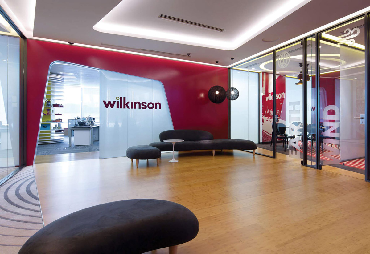 Aedas Interiors’ Wilkinson Asia Headquarters in Hong Kong is shown with a brand‑forward collaborative environment featuring a curved white innovation room, bold red feature walls, timber flooring, and glazed meeting suites. The image highlights flexible lounge seating, strong corporate branding integration, and spatial planning that prioritises informal collaboration and dynamic interaction across the workplace.