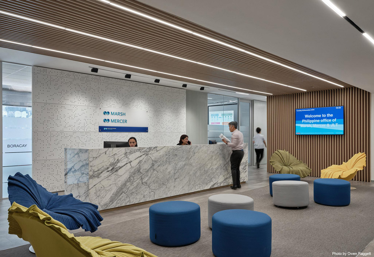 Aedas Interiors’ Marsh & McLennan Manila office is shown with a formal reception zone featuring a marble‑clad counter, vertically slatted timber wall, integrated digital signage, and open‑plan seating. The image highlights brand‑focused interior architecture, acoustic detailing, and a flexible workplace designed to support a fully open, non‑assigned work environment during organisational transition.