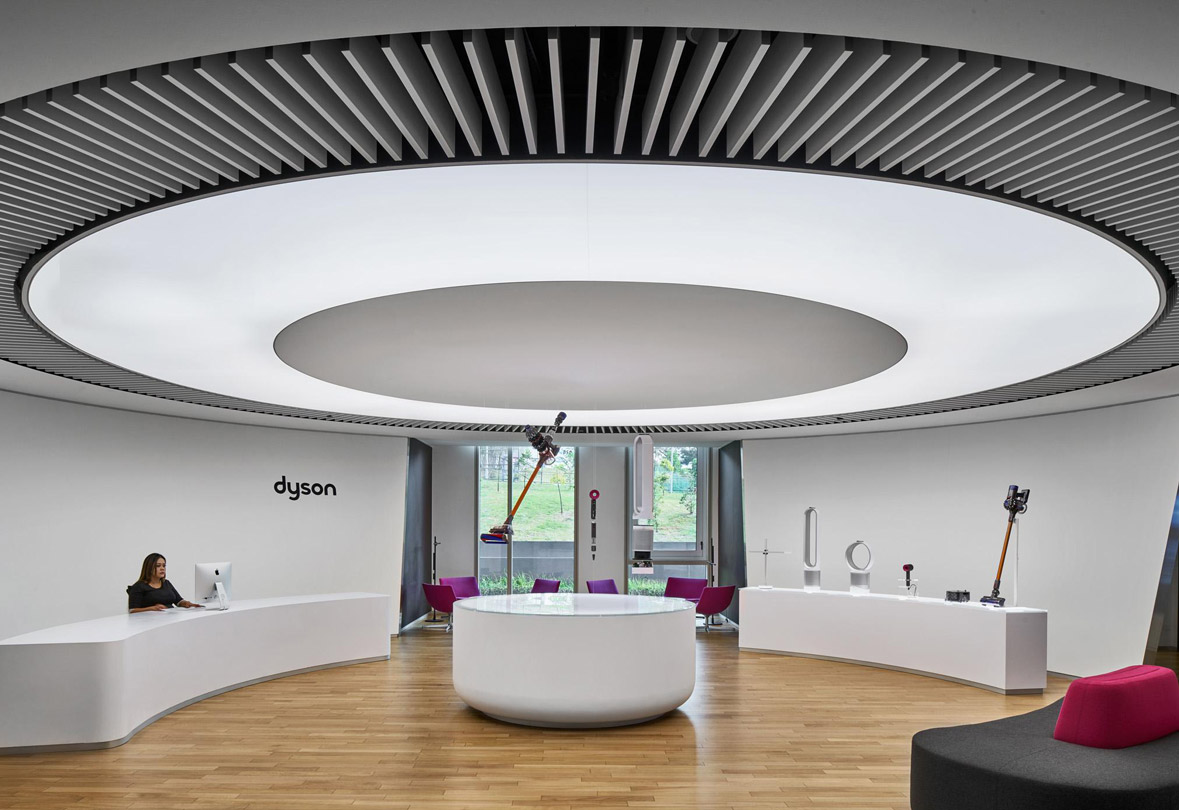 Aedas Interiors’ Dyson Singapore Technology Centre is shown as a circular collaboration and display space defined by a large ring‑shaped ceiling light, curved white demonstration counters, and timber flooring. The image highlights a technology‑led interior with integrated product showcases, central gathering focus, and spatial planning that supports activity‑based working and collaborative engineering culture.